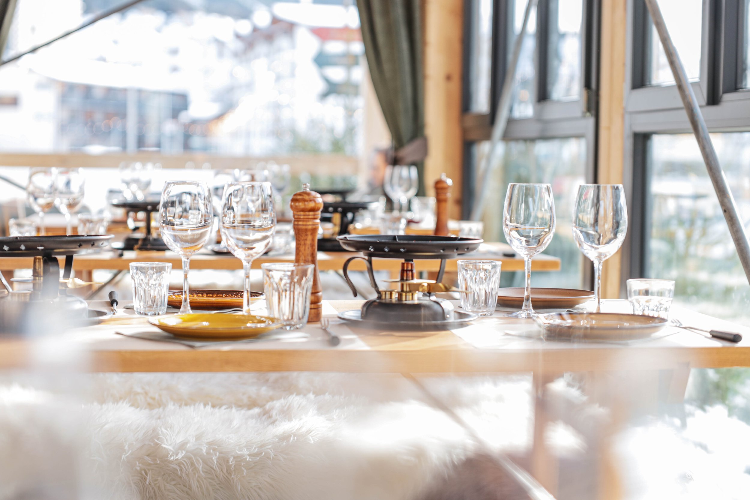 Ein festlich gedeckter Holztisch mit Weingläsern, Wasserkrügen, Salz- und Pfefferstreuern, Tellern und Besteck in einem hell erleuchteten Raum mit großen Fenstern und Blick nach draußen.