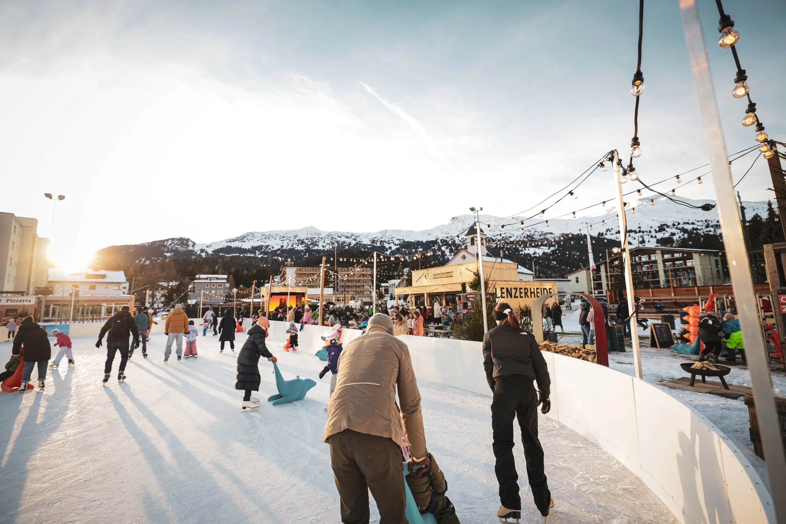 Menschen beim Schlittschuhlaufen auf einer Ice Rink mit Bergkulisse im Hintergrund, Sonnenuntergang und Lichterketten.