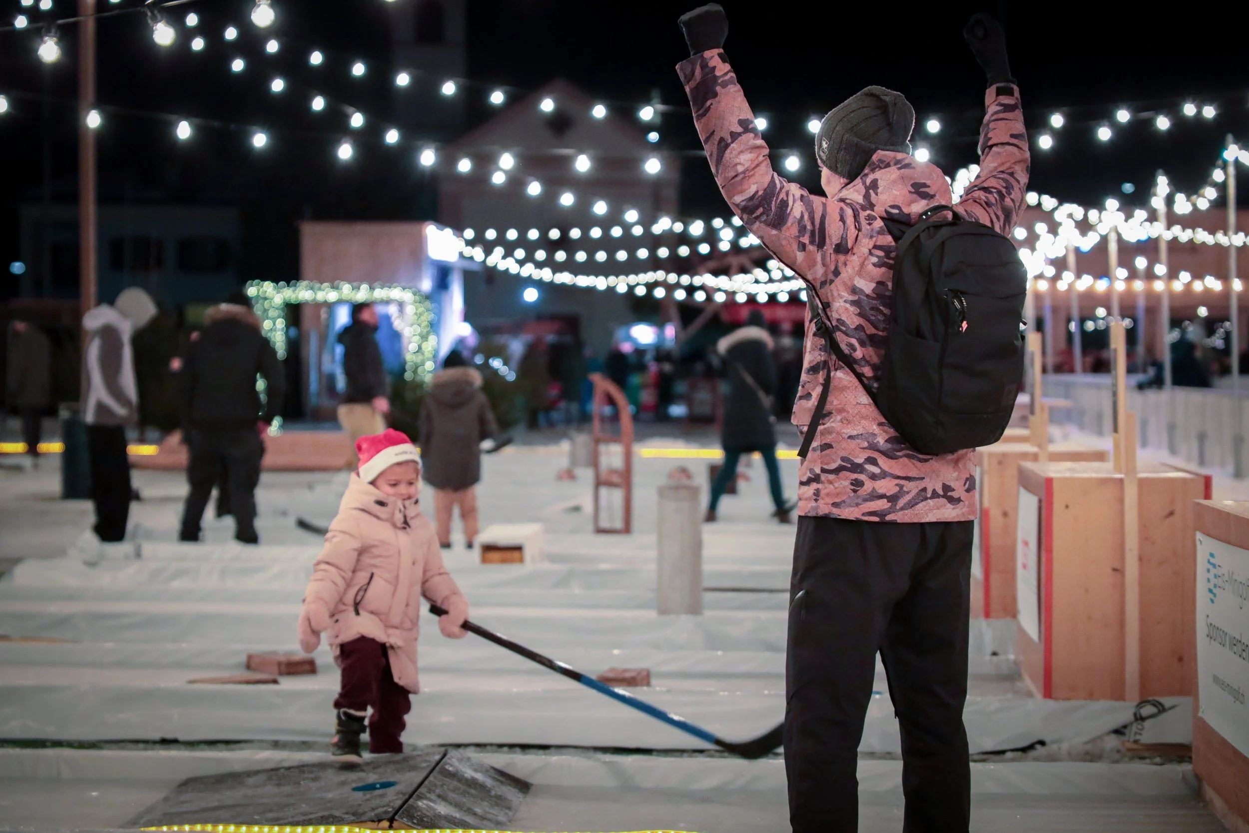 Ein Junge in einem pinken Wintermantel und Mütze spielt mit einem Hockeyschläger auf einer Eisbahn, während ein erwachsener Begleiter mit verschränkten Armen zuschaut. Im Hintergrund sind weitere Personen, Lichter und weihnachtliche Dekorationen bei Nacht.