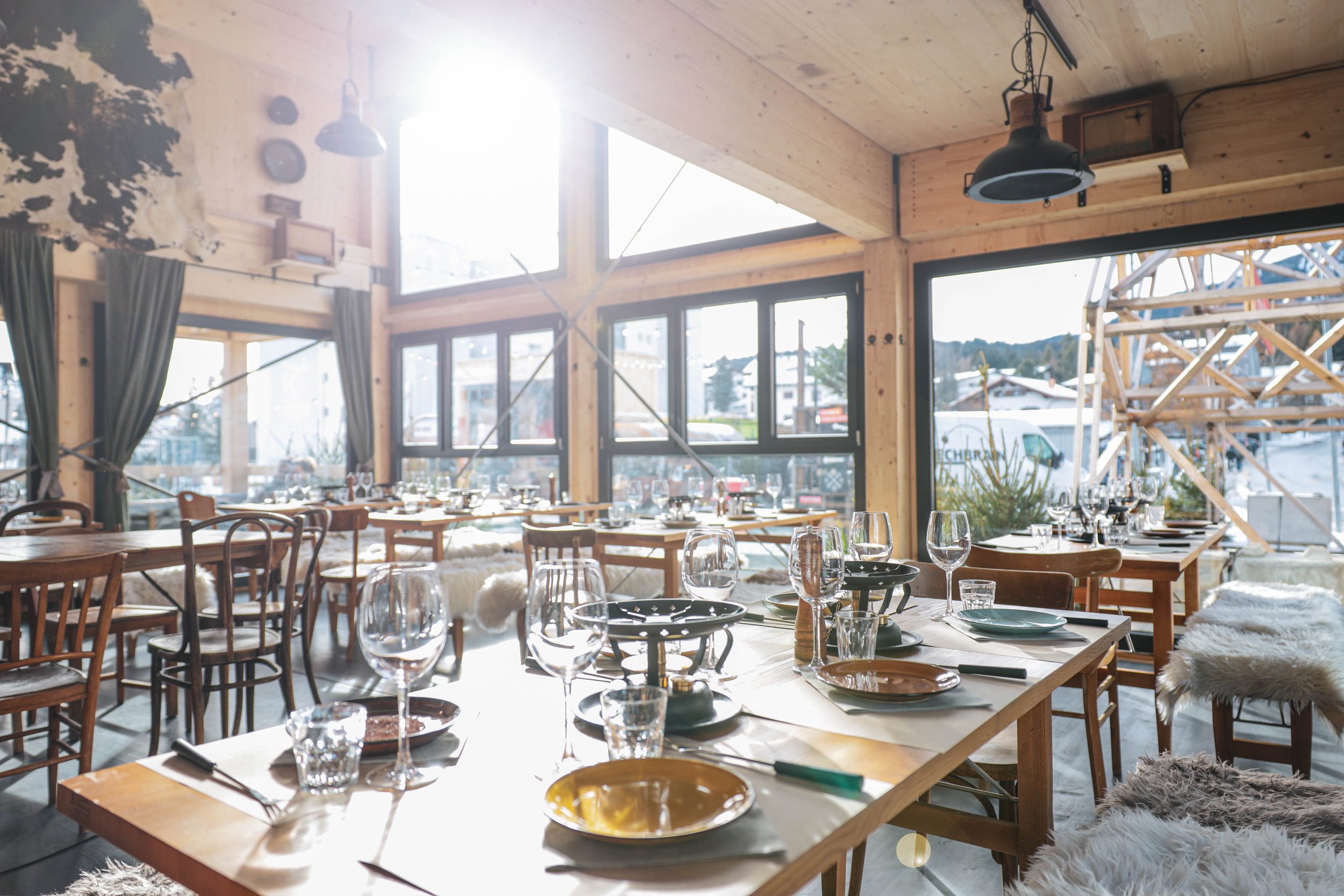 Ein gemütliches, helles Restaurant mit viel Holzdekoration und großen Fenstern, gedeckter Tisch mit Gläsern, Tellern und Besteck, winterliche Dekoration und Aussicht auf Schnee im Außenbereich.