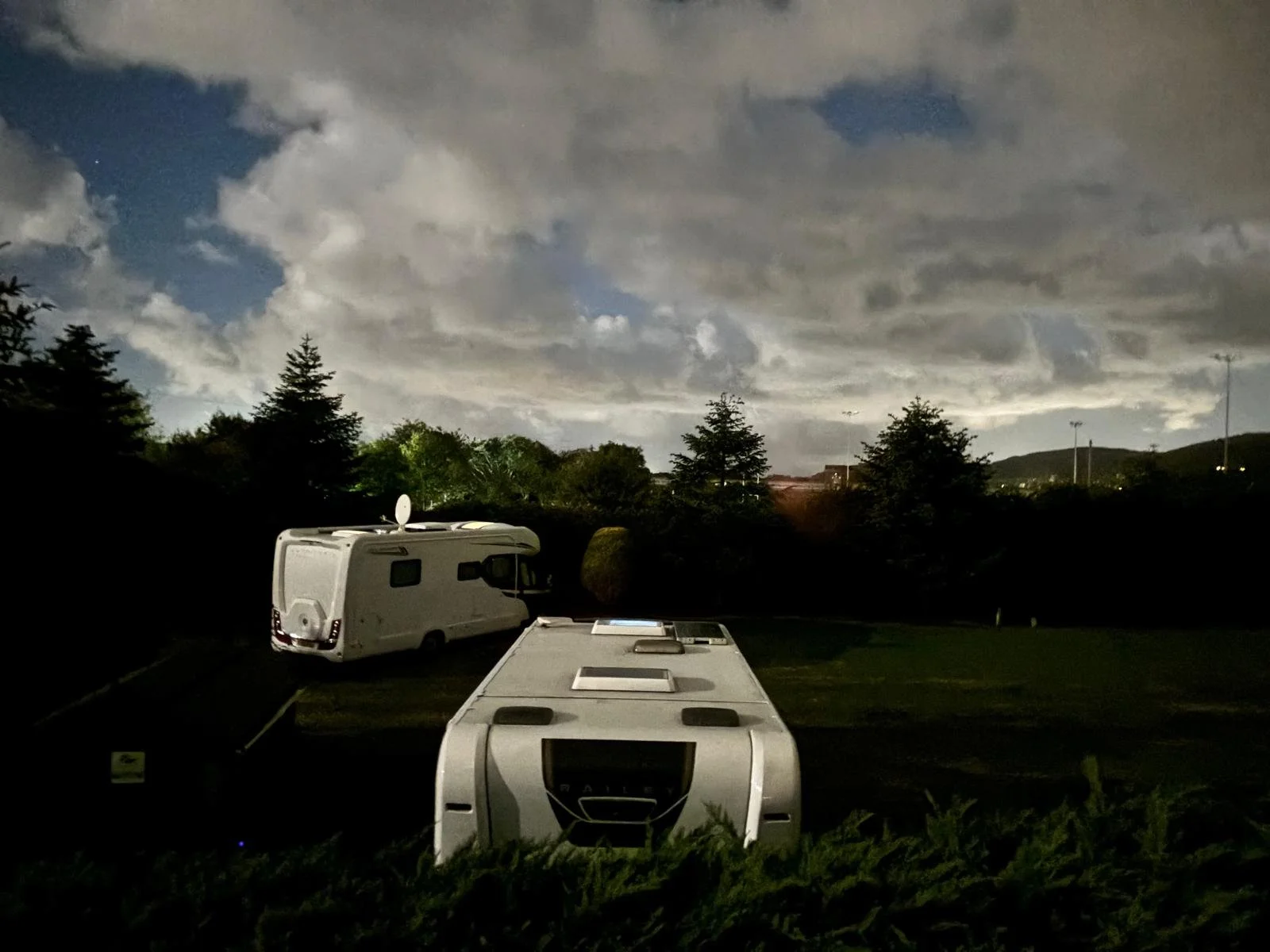 Nighttime view of two parked caravans in a grassy area, with a cloudy sky and dark trees in the background.
