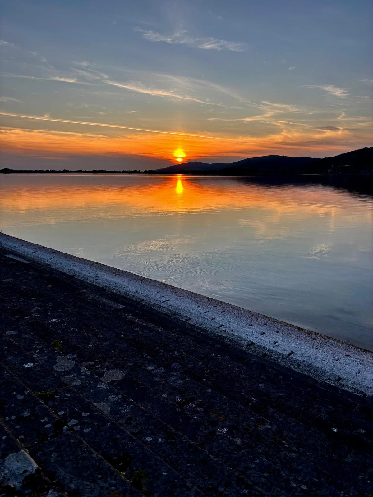Sunset over a body of water with mountains in the background, calm water reflecting the sky, and a dark foreground of a stone or concrete edge.