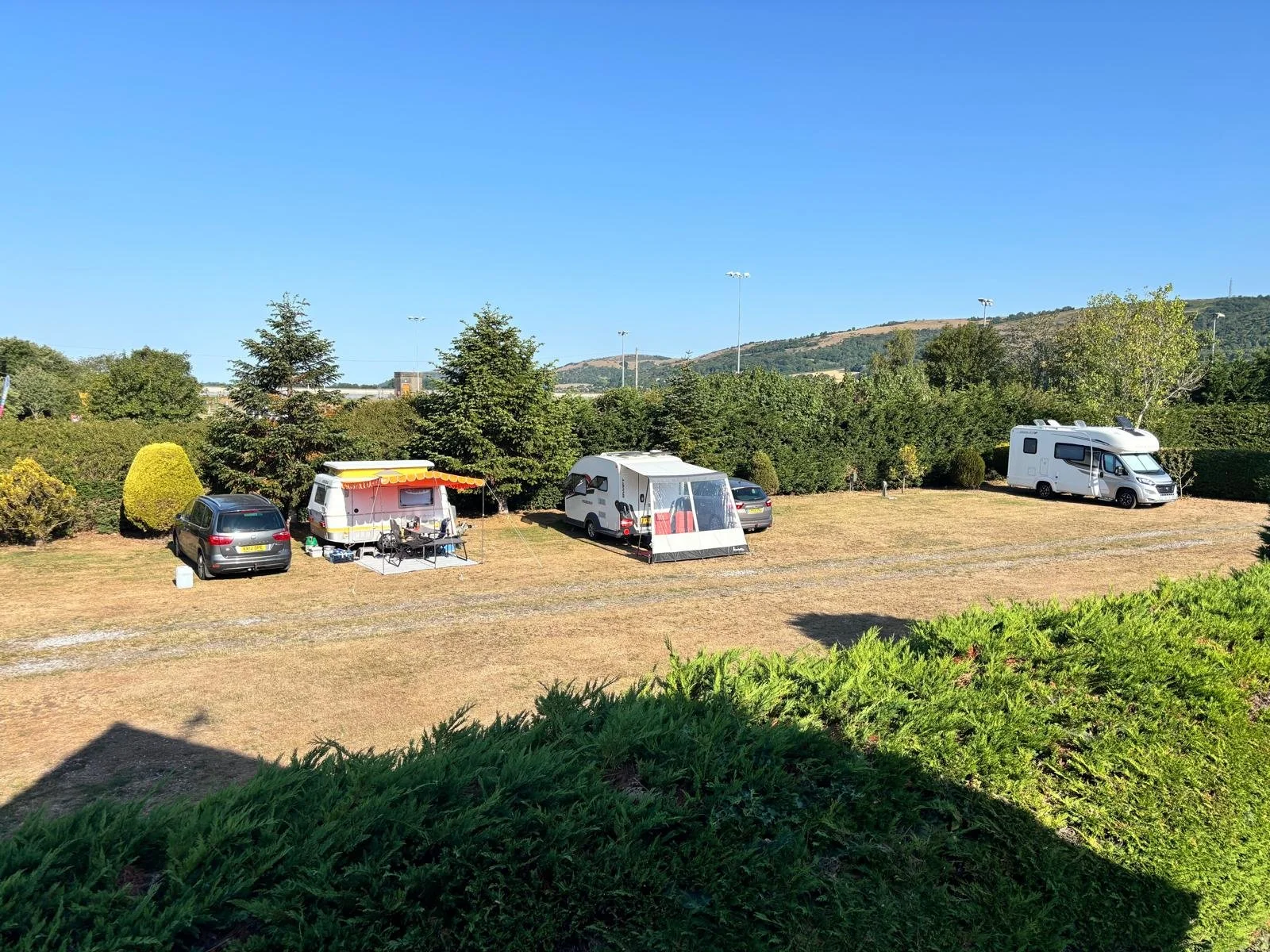 A scenic view of a campground with several parked vehicles, including a camper trailer, a caravan with a tent, a motorhome, and a car, set among green trees under a clear blue sky.