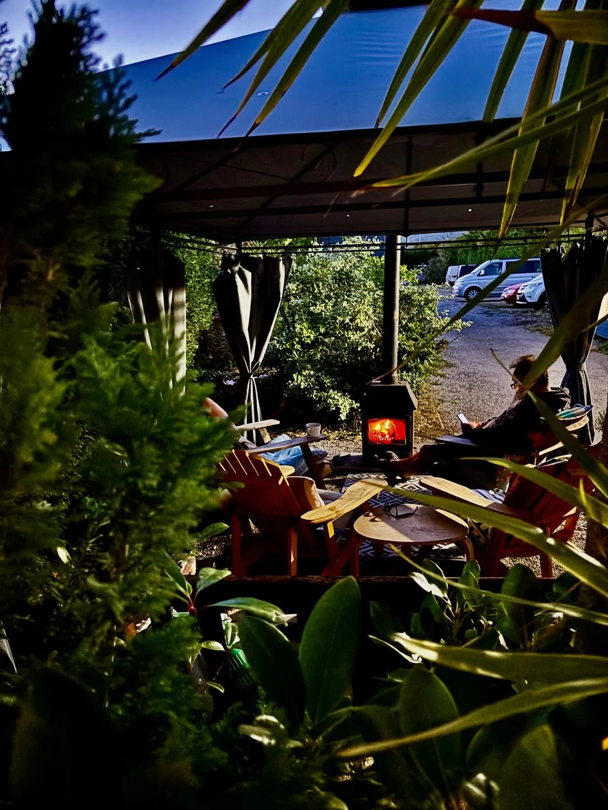Outdoor seating area at night with a small heater, surrounded by lush green plants and trees, and a person sitting using a phone. Car parking lot visible in the background.