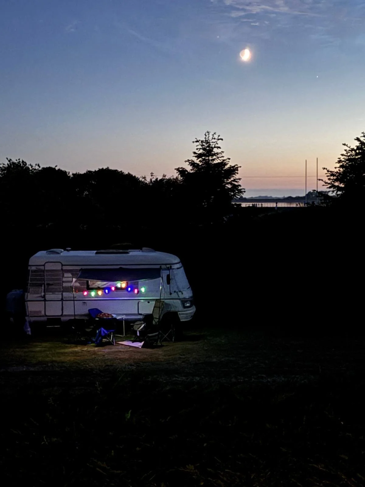 A camper trailer decorated with colorful string lights sits on a grassy area at dusk, under a sky with a bright crescent moon and a few stars, with trees and a body of water in the background.
