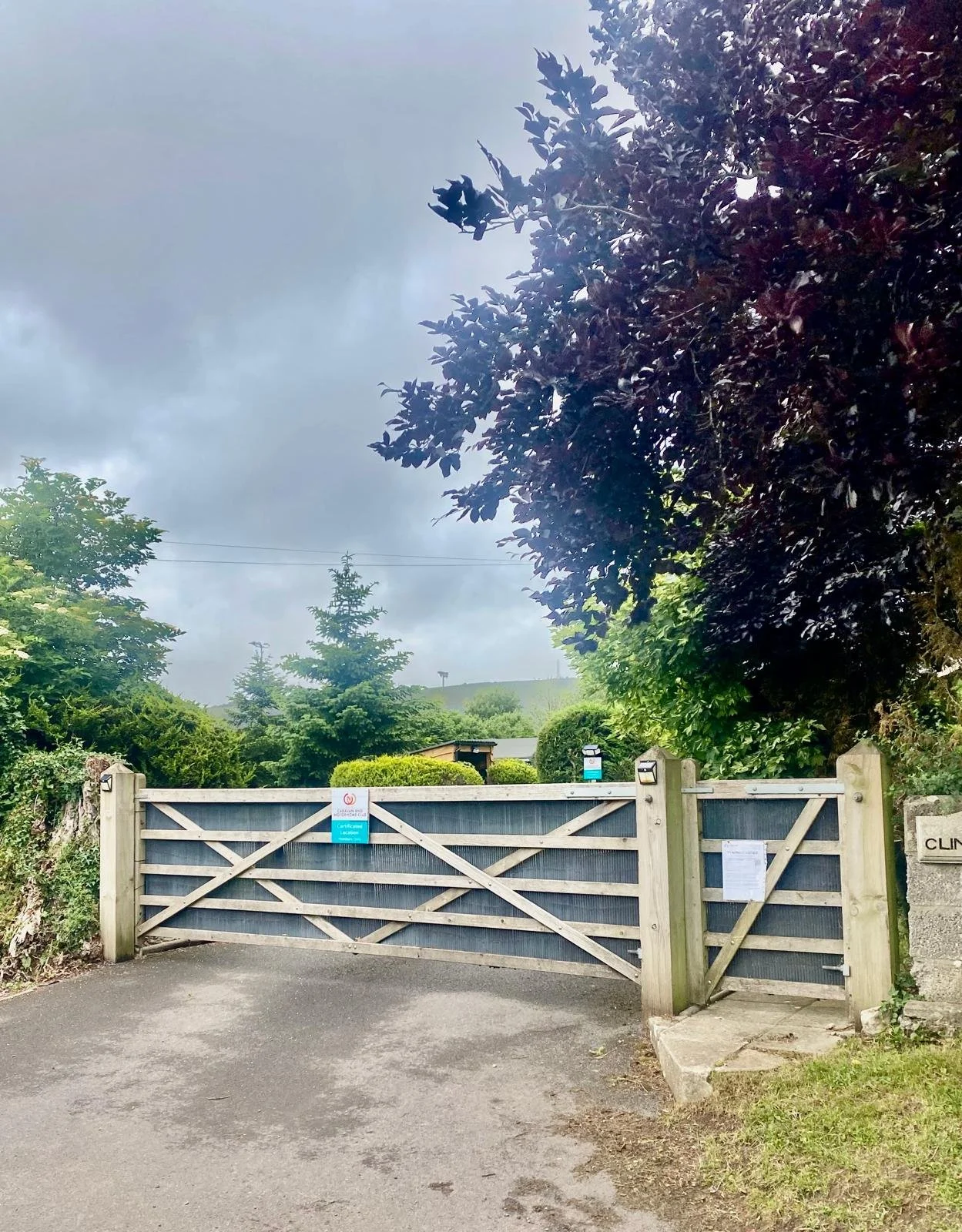 A wooden gate with blue panels blocking the entrance to a property, surrounded by lush green trees and shrubs, with cloudy skies overhead.