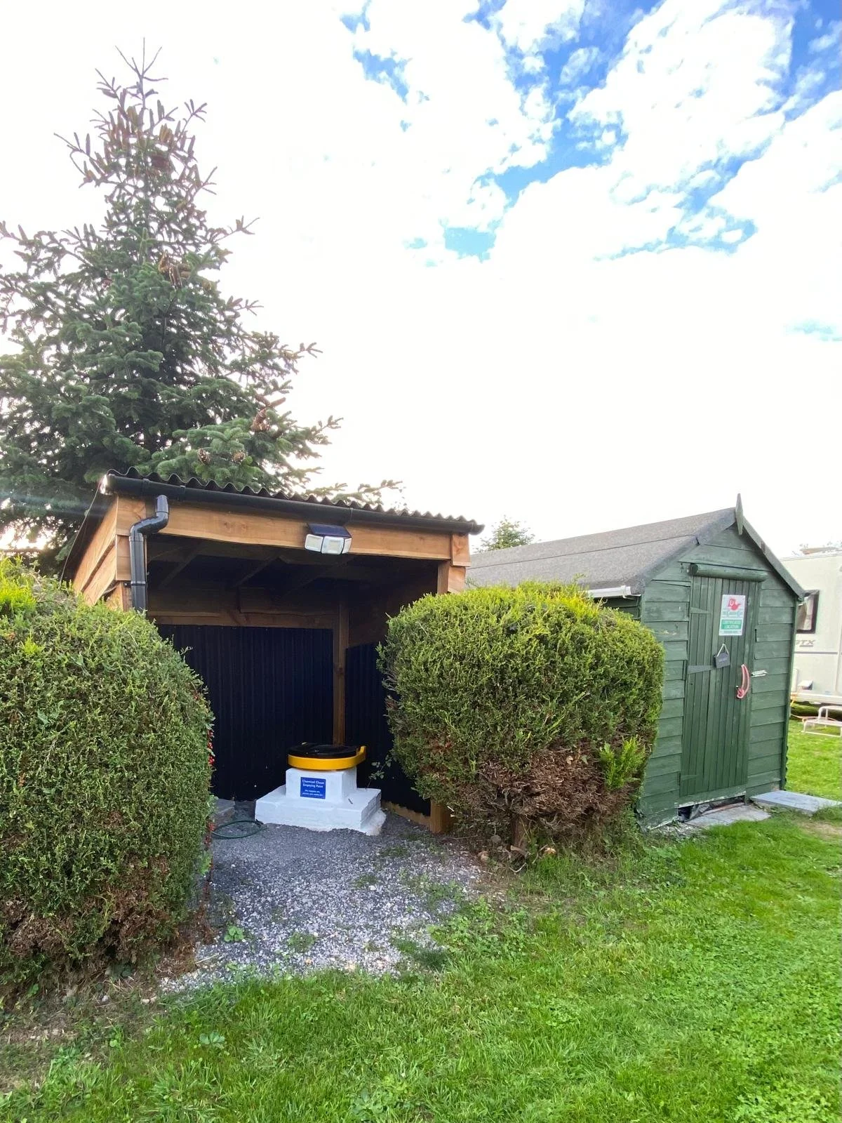 A small wooden outdoor shelter with a sloped roof, situated next to a green storage shed, surrounded by bushes and grass, under a partly cloudy sky.