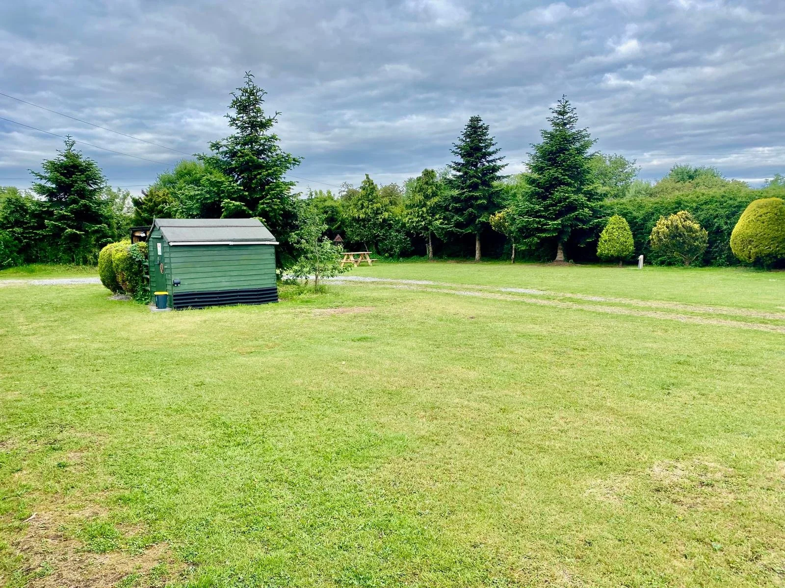 Open grassy field with a small green shed on the left, several trees in the background, and overcast clouds overhead.