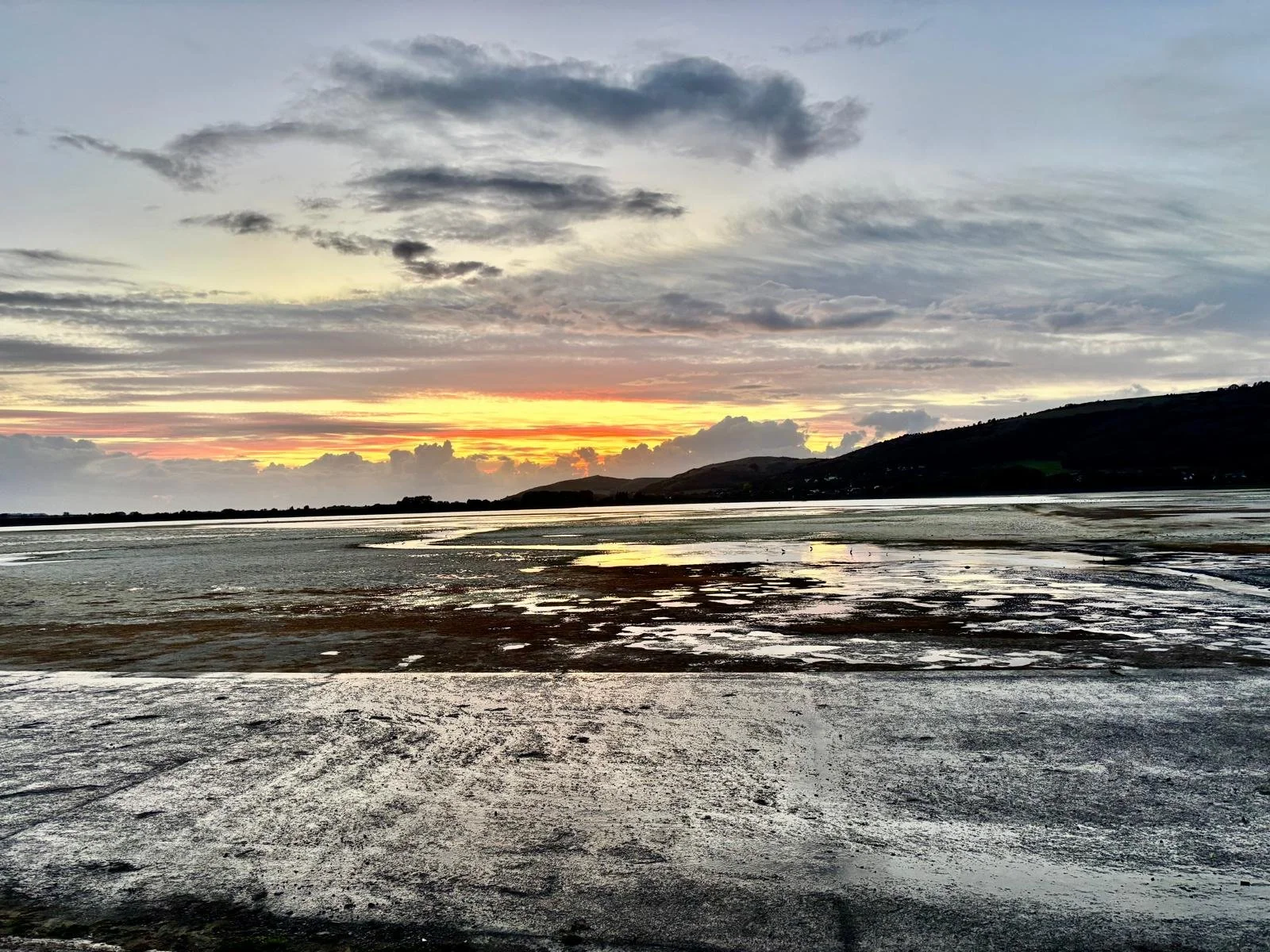 Scenic view of a beach or shoreline at sunset with a colorful sky, clouds, distant hills, and reflections on wet sand.