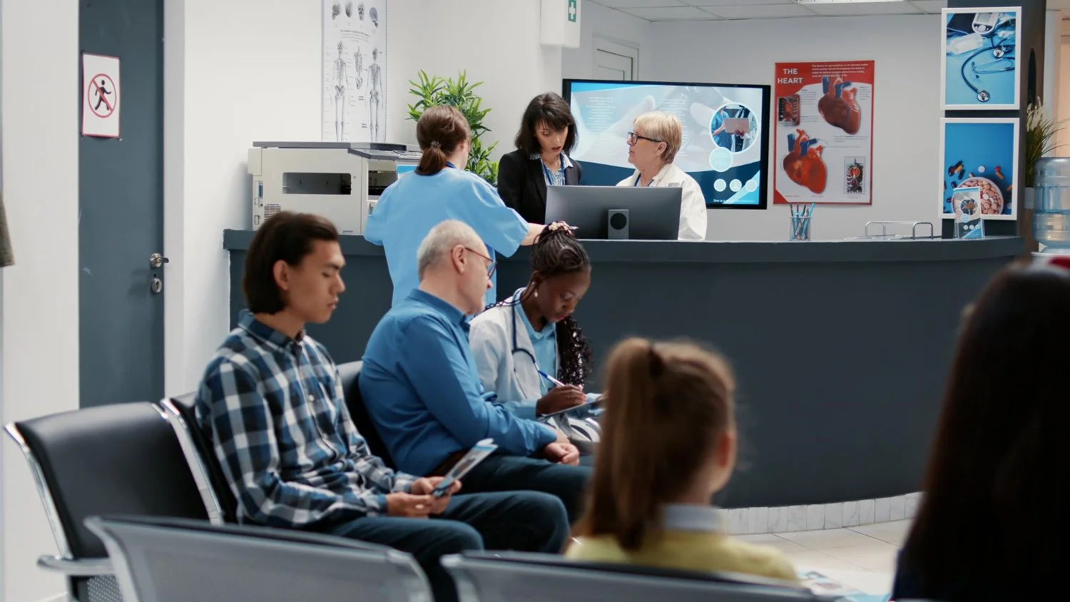 People sitting in a waiting area of a hospital or clinic, with a reception desk in the background where medical staff are talking and working.