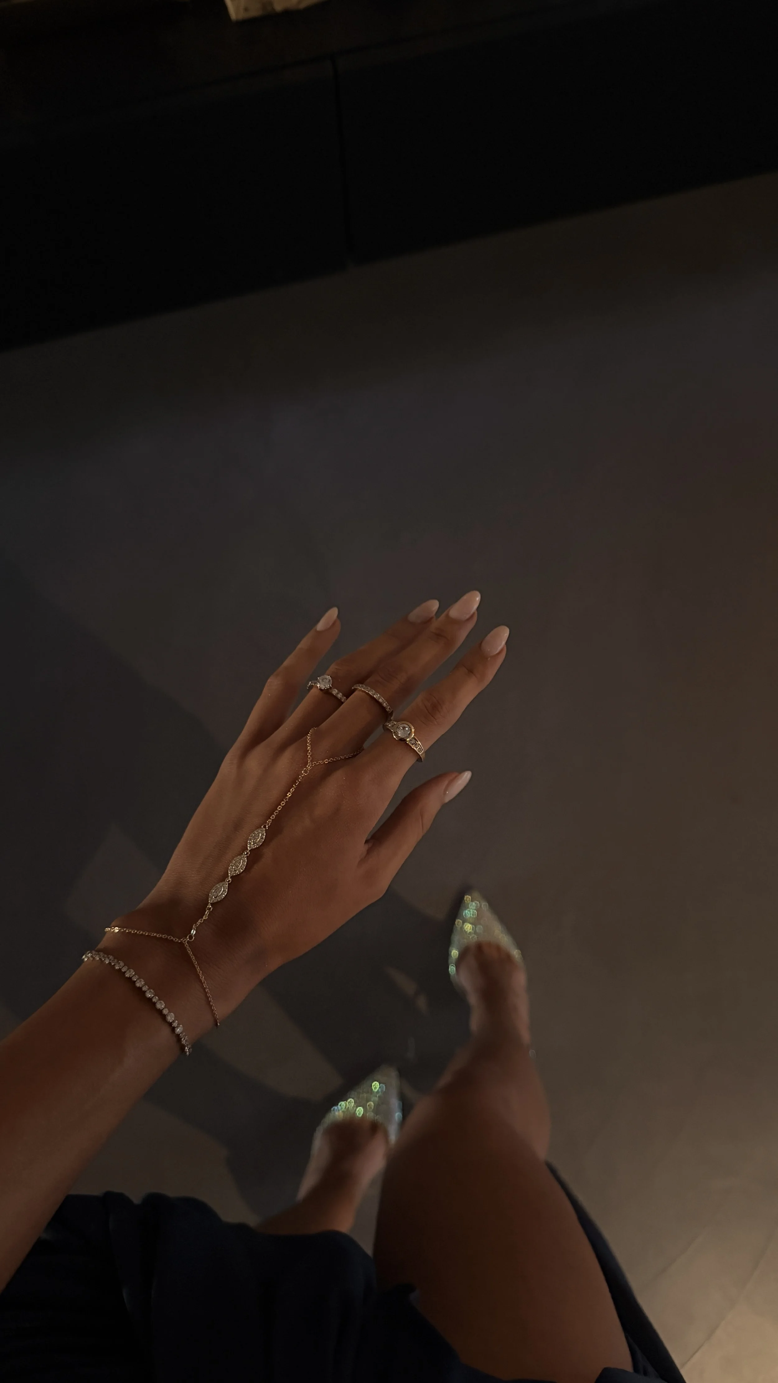 A hand with multiple rings and a bracelet, displaying a wrist with a delicate chain bracelet, and wearing sparkly high-heeled shoes.