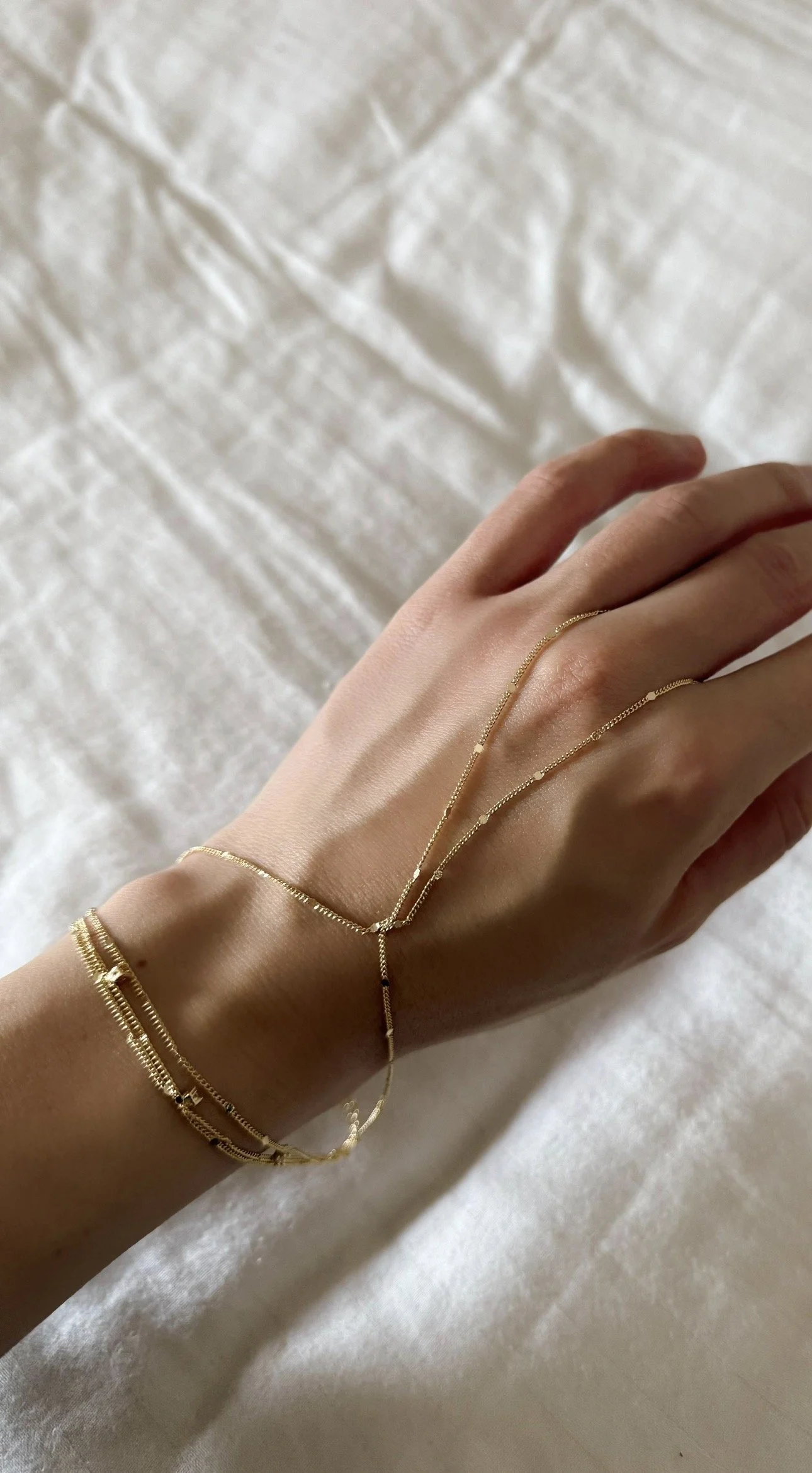 Multiple gold bracelets and a gold chain bracelet on a person's wrist and hand resting on a light-colored fabric background.