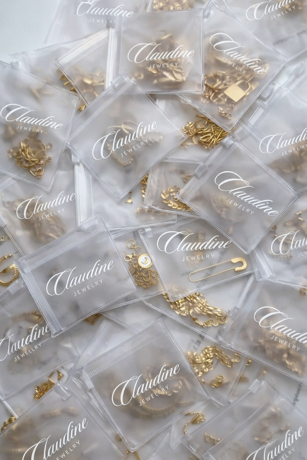 Multiple small transparent plastic bags with the brand name 'Claudine Jewelry' printed on them, containing gold jewelry pieces such as chains, charms, and pins.