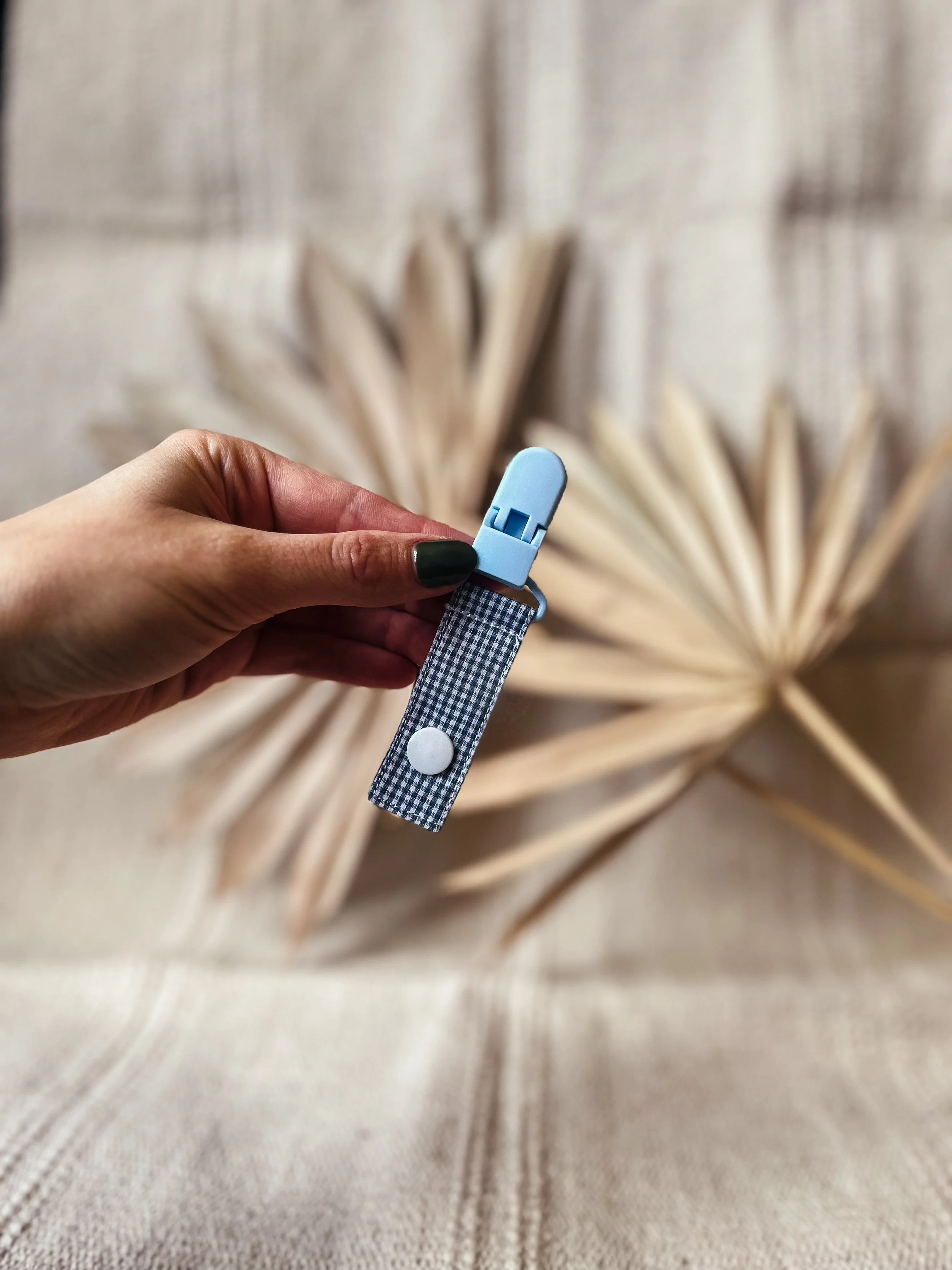 Eine Hand hält einen blauen, clipartigen Gegenstand mit einem Stoffband, vor einem Hintergrund aus getrockneten Palmblättern.