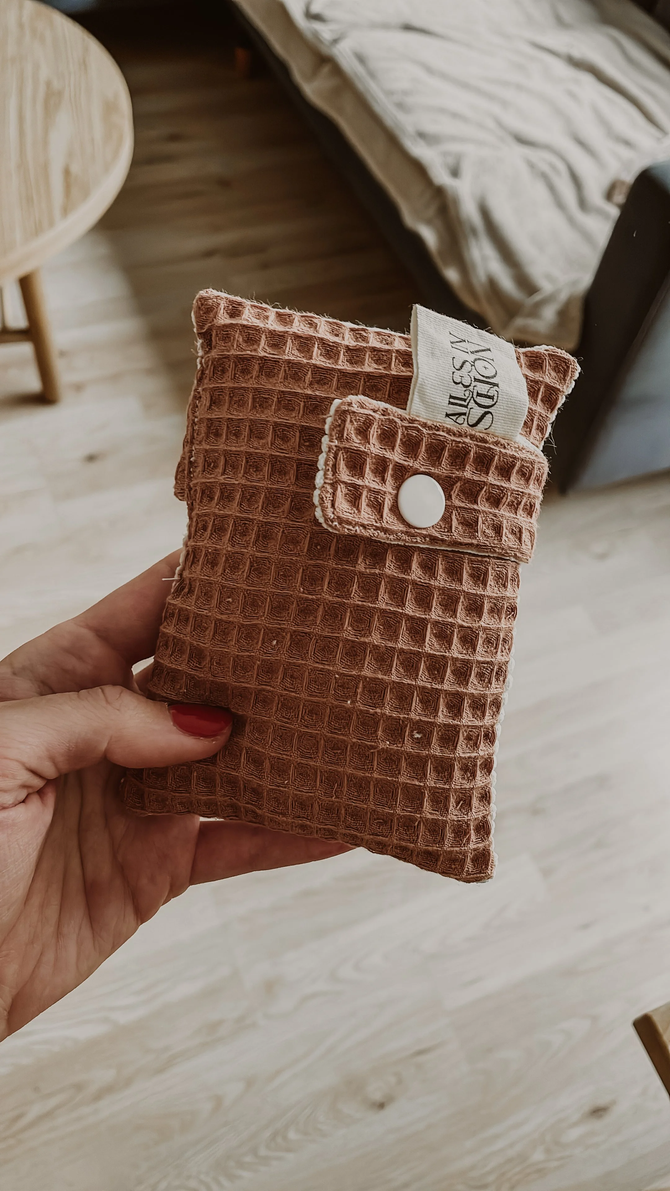 Eine Hand hält eine grob strukturierte, rosafarbene Waschlappen mit Knopf und Wäscheklammer, im Hintergrund ein Bett und ein Holztisch.