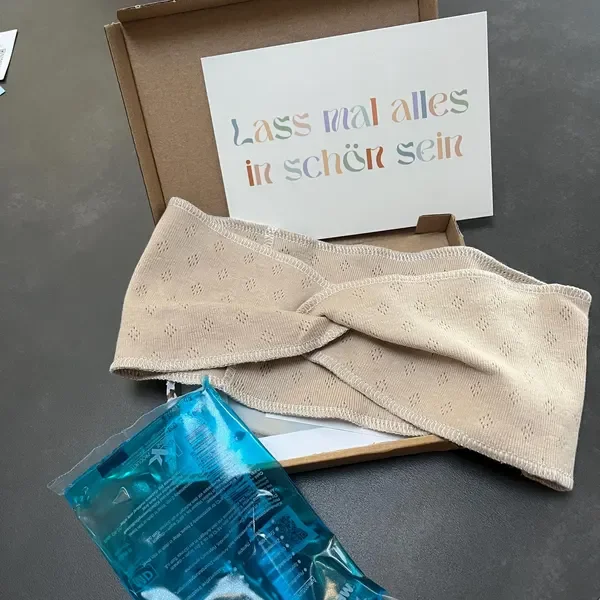 Ein beige Kopfband, eine blaue Wasserflasche, eine weiße Karte mit buntem deutschen Text, die in einem braunen Karton liegt.
