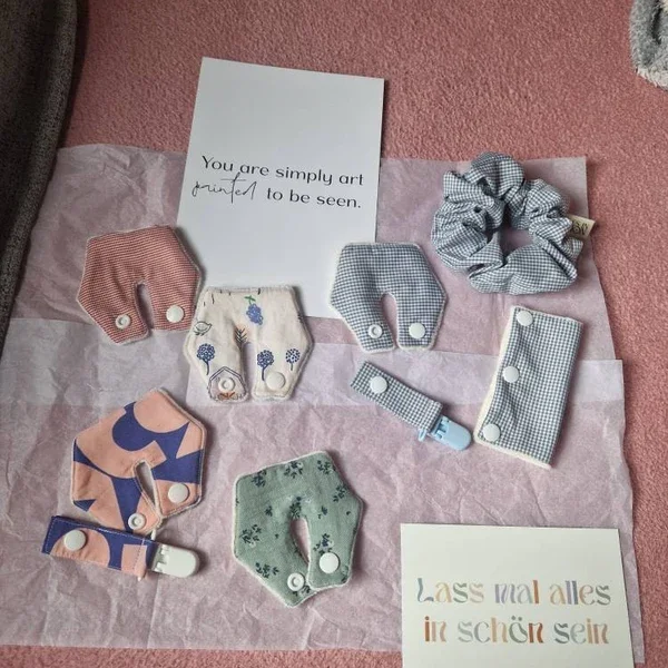 Verschiedene gewebte Haargummis und Haarspangen in Pastellfarben, auf einem rosa Papieruntergrund, mit zwei ausgeschnittenen Sprüchen, die positive Botschaften enthalten.