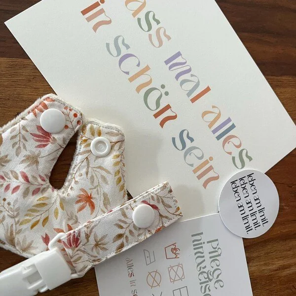 Ein Stoffband mit Druckknöpfen, eine Broschüre mit buntem Text und einem runden Aufkleber mit deutschem Text liegen auf einem Holztisch.