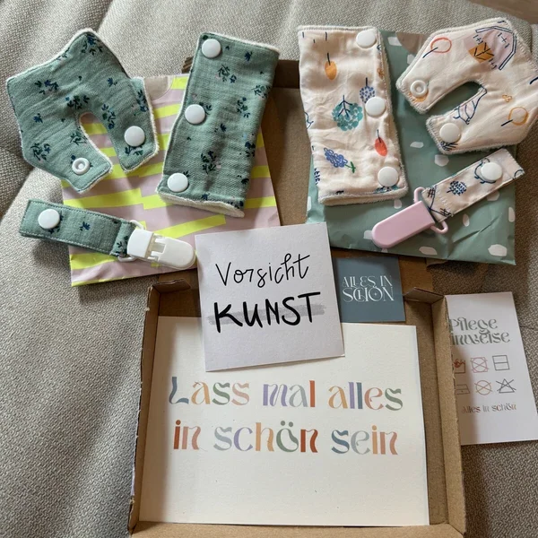 Mehrere Stoffbinden und schützende Textilien mit unterschiedlichen Mustern auf einem Tisch, begleitet von einem handgeschriebenen Zettel mit der Aufschrift 'Vorsicht KUNST' und einem weiteren Zettel mit der Botschaft 'Lass mal alles in schön sein'.