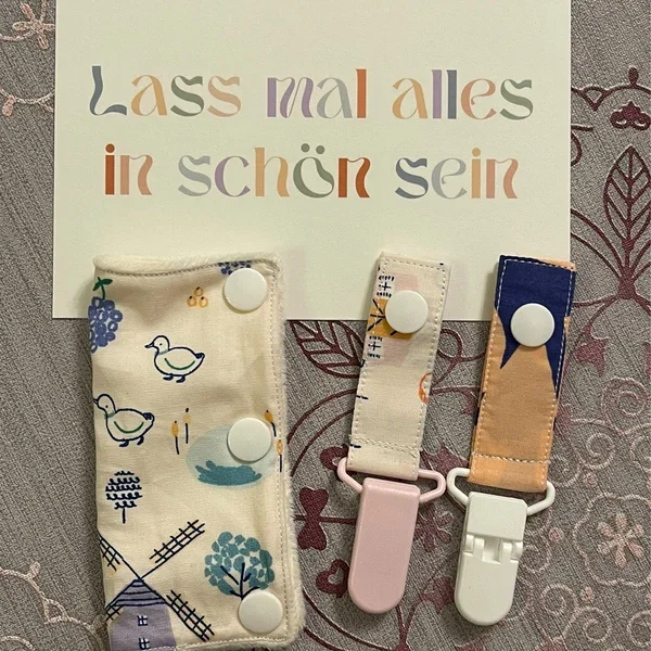 Eine Karte mit dem deutschen Satz 'Lass mal alles in schön sein', umgeben von drei Schnulleranhängern auf einem Stickstofftuch.