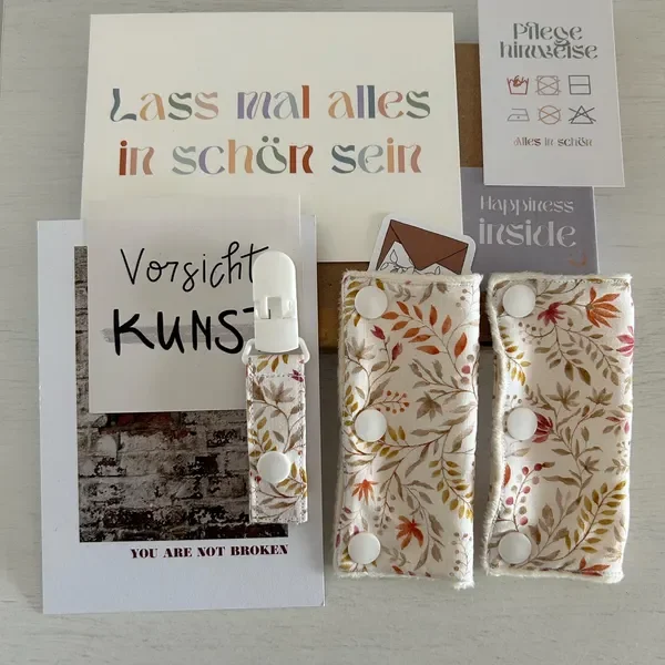 Verschiedene Papier- und Text-Prints, darunter ein Zitat 'Lass mal alles in schön sein', eine Handtasche, zwei Stoffbeutel mit Druckknöpfen, eine Packung Pflegehinweise, und ein kleiner Zettel 'Vorsicht Kunst', alles arrangiert auf einem weißen Tisch.