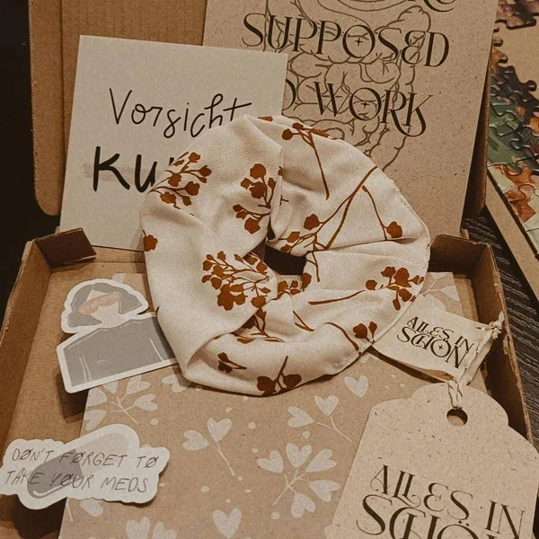 Ein aufklappbares Geschenkset mit einer Scrunchie, einem Handtuch mit der Aufschrift 'Alles in Ordnung', und verschiedenen Karten, darunter eine mit 'Vorsicht Kuschelzeit' und eine andere mit 'Supposed to work'.