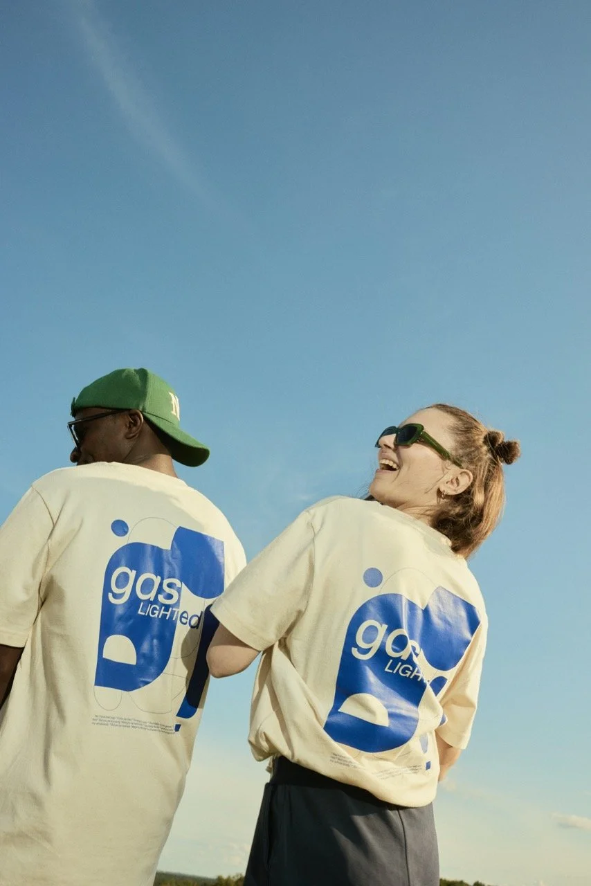 Zwei lachende Personen tragen off-white T-Shirts mit einem blauen Design. Sie stehen im Freien unter einem blauen Himmel. Einer trägt einen grünen Basecap, beide sehen entspannt und fröhlich aus.