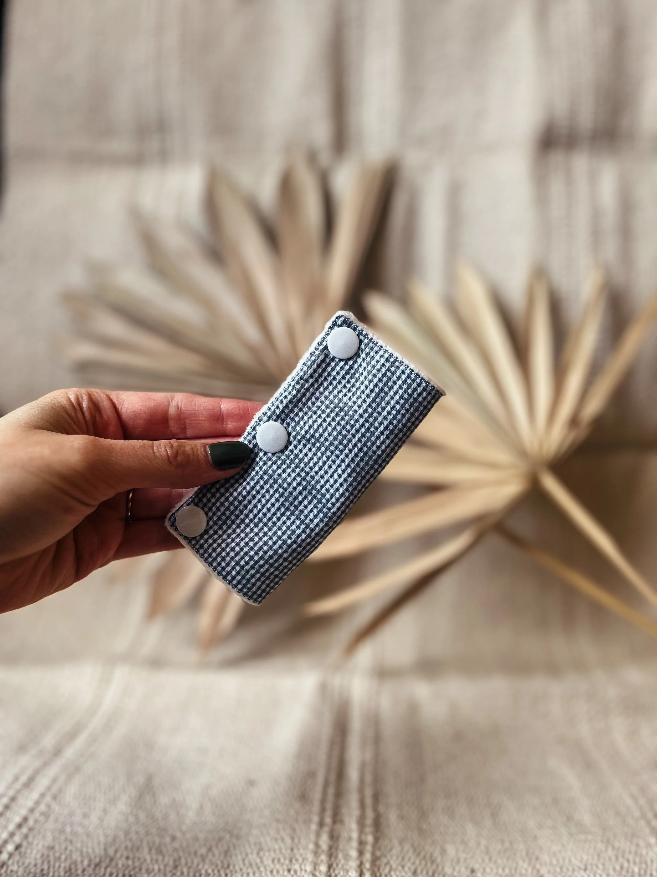 Hand hält eine gecheckte Brillenetui vor einem Hintergrund aus getrockneten Palmenblättern.