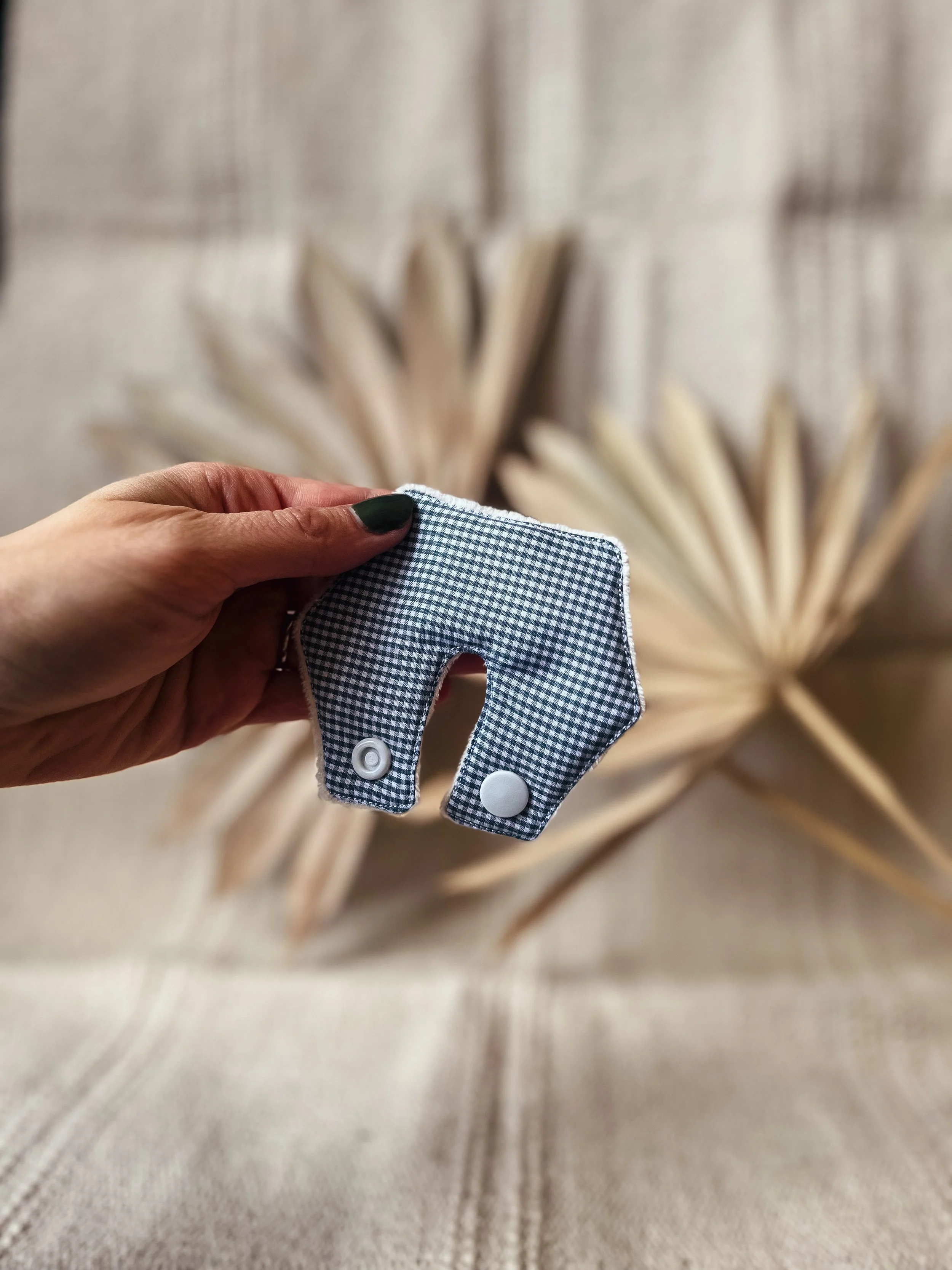 Hand hält ein Stoff-Accessoire in Form eines Babys, das aus grau-weiß kariertem Stoff besteht, vor zwei großen, getrockneten Palmblättern im Hintergrund.