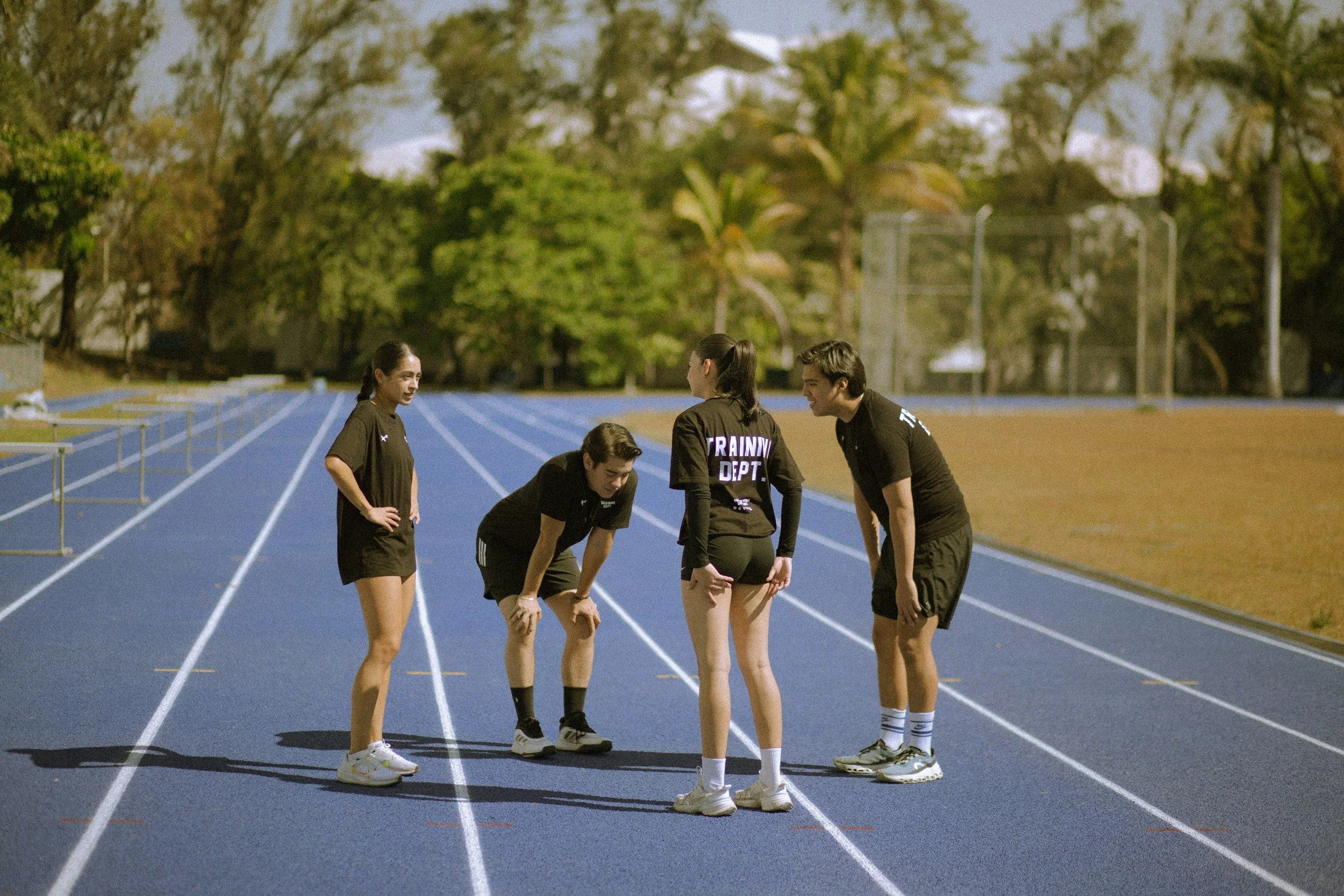 Four teenagers in athletic clothing standing on a running track, with some bending over and one with the words 'RAINY DEPT.' on the back, surrounded by trees in the background.