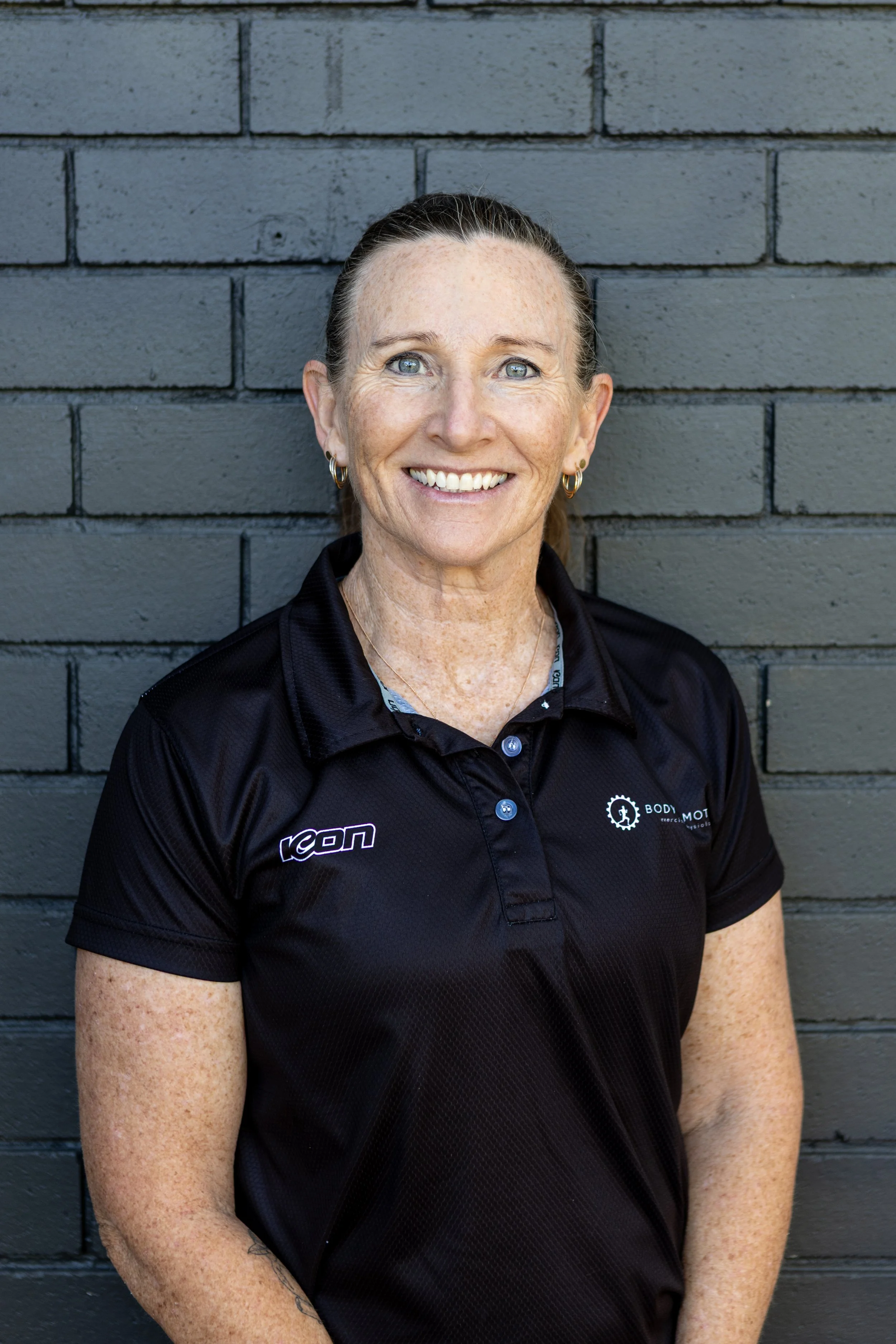 Woman with brown hair tied back, smiling, wearing a black sports shirt with logos, standing against a gray brick wall.