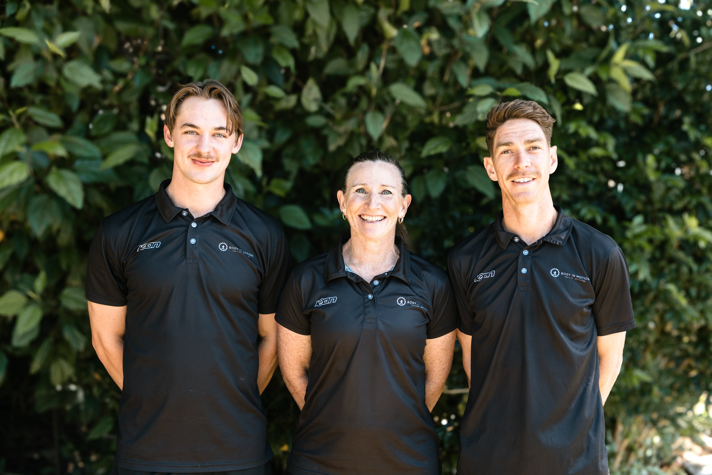 Three people standing outdoors in front of green foliage, wearing black polo shirts with 'Body in Motion Exercise Physiology' logos, smiling at the camera.