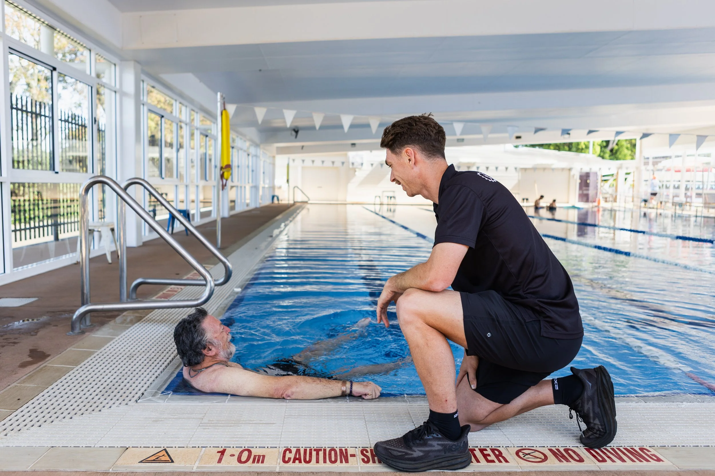 Exercise physiologist assisting a man during swim training in an indoor pool.
