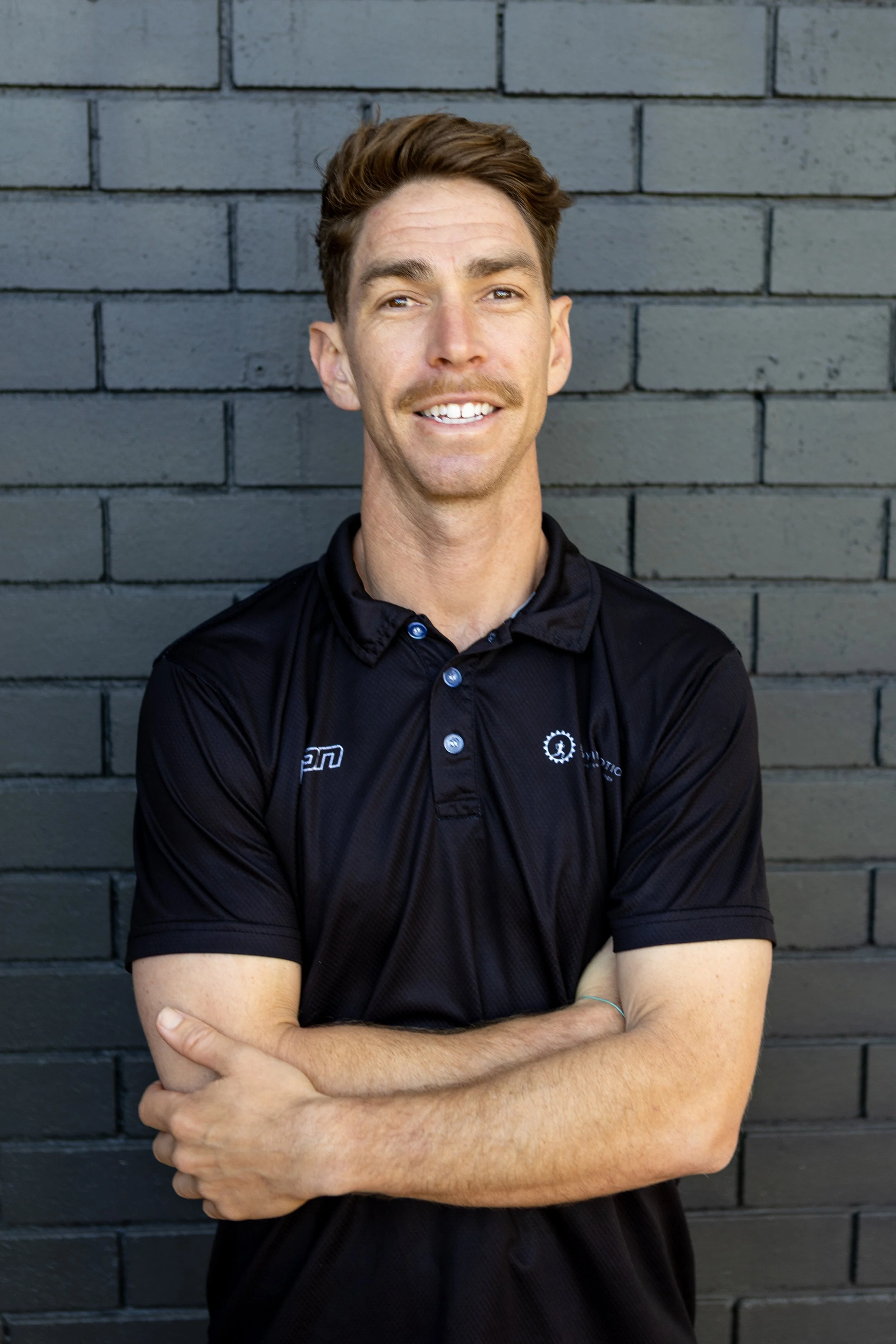 A young man with short brown hair, light facial hair, and a slight smile, standing against a black brick wall, wearing a black polo shirt with logos.