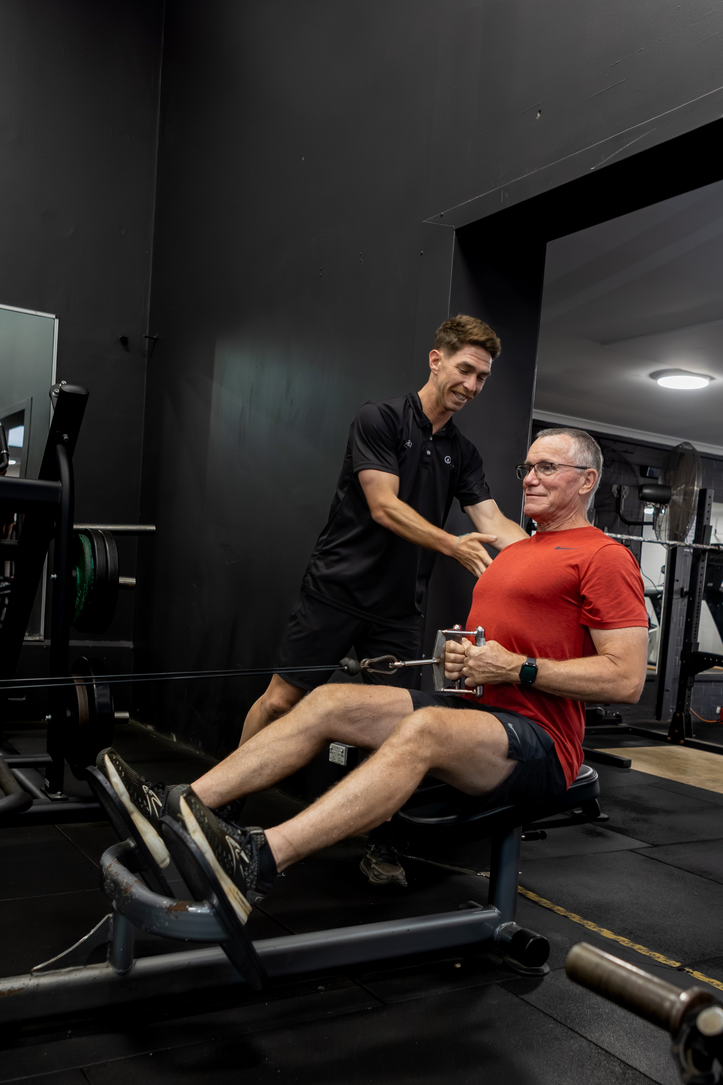 An exercise physiologist assisting an older man with a seated cable exercise in a gym. The man is wearing a red shirt and black shorts, while the exercise physiologist is wearing a black shirt and shorts. Both are smiling and engaged in the workout.