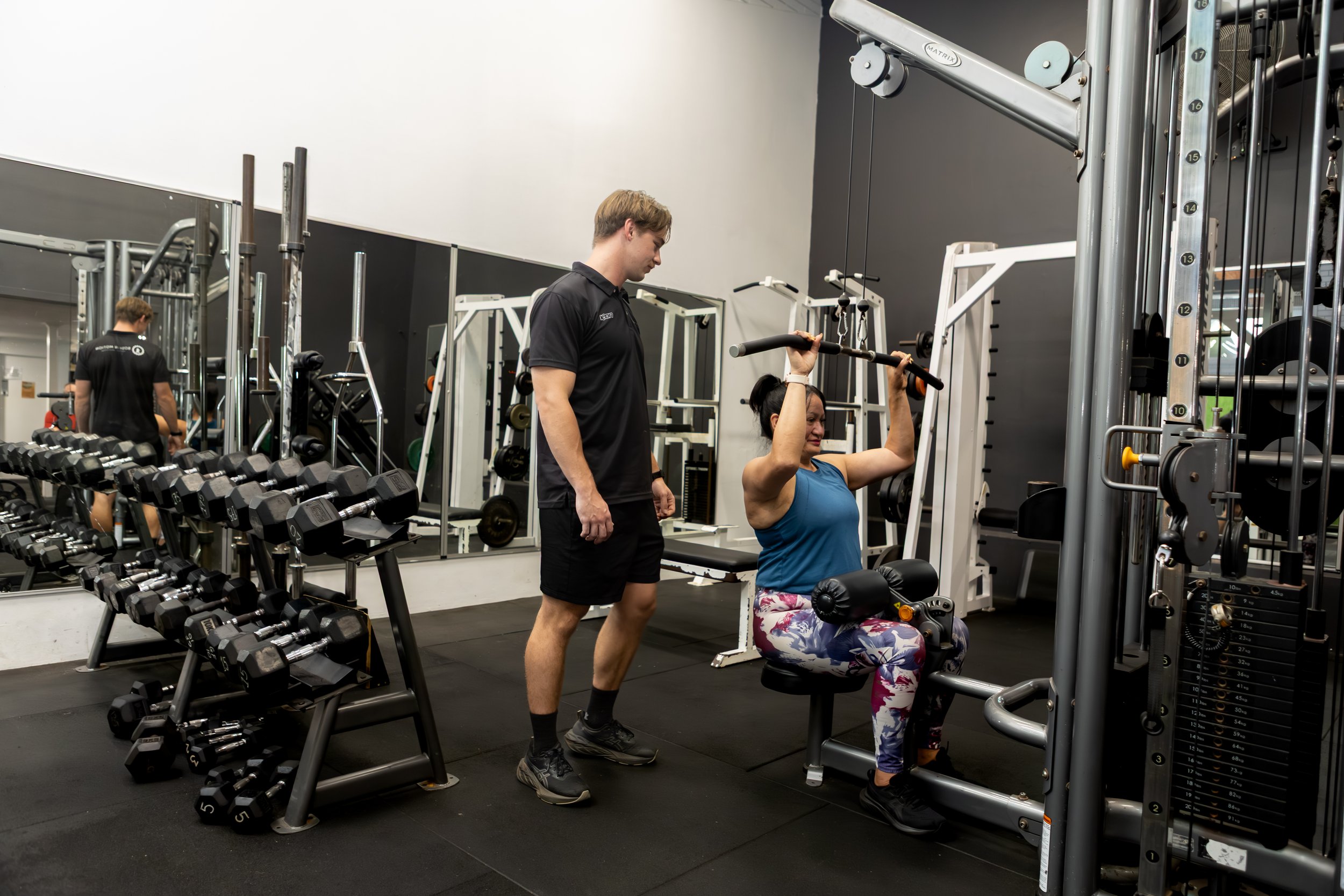 A woman in blue top and colorful leggings performs a shoulder press on a machine while an exercise physiologist watches on in a gym.