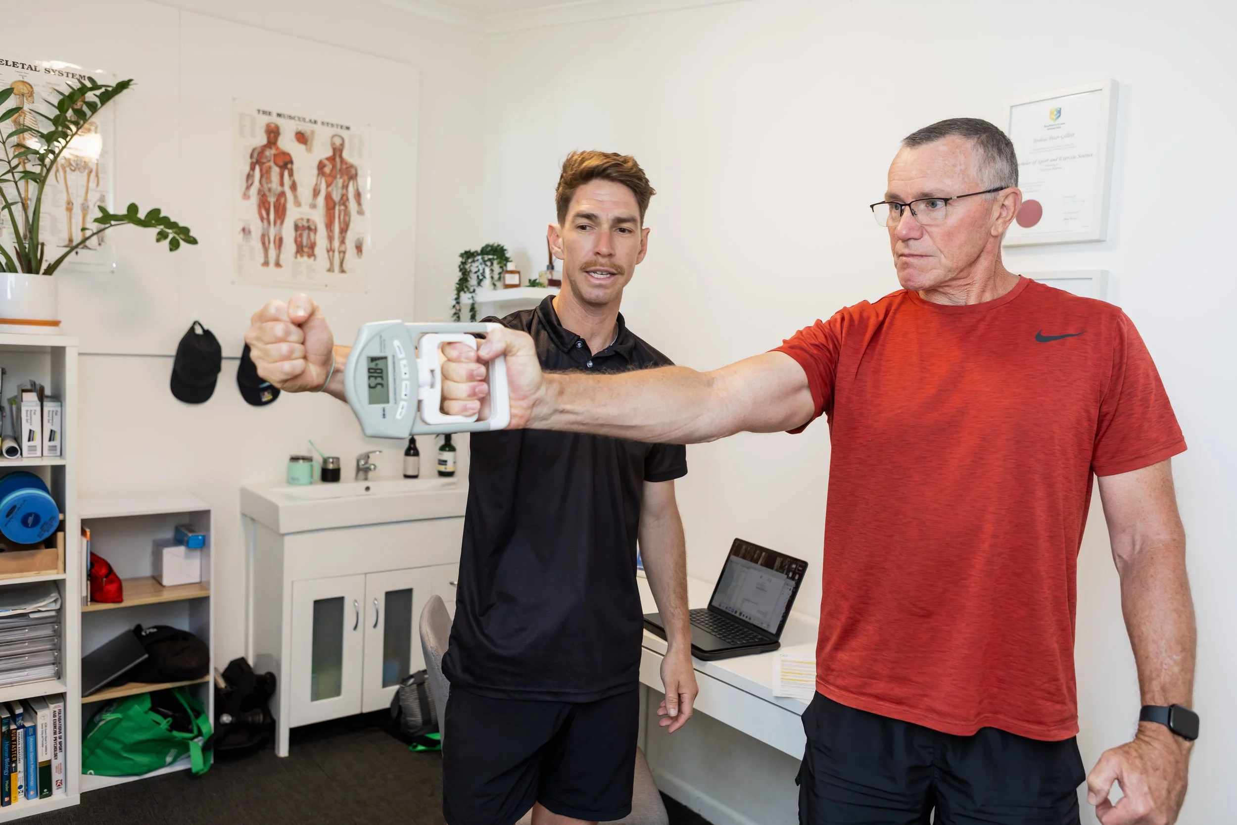 An older man in a red athletic shirt and glasses is performing a resistance arm exercise with a handheld device, supervised by an exercise physiologist with brown hair in a black shirt, in an office.