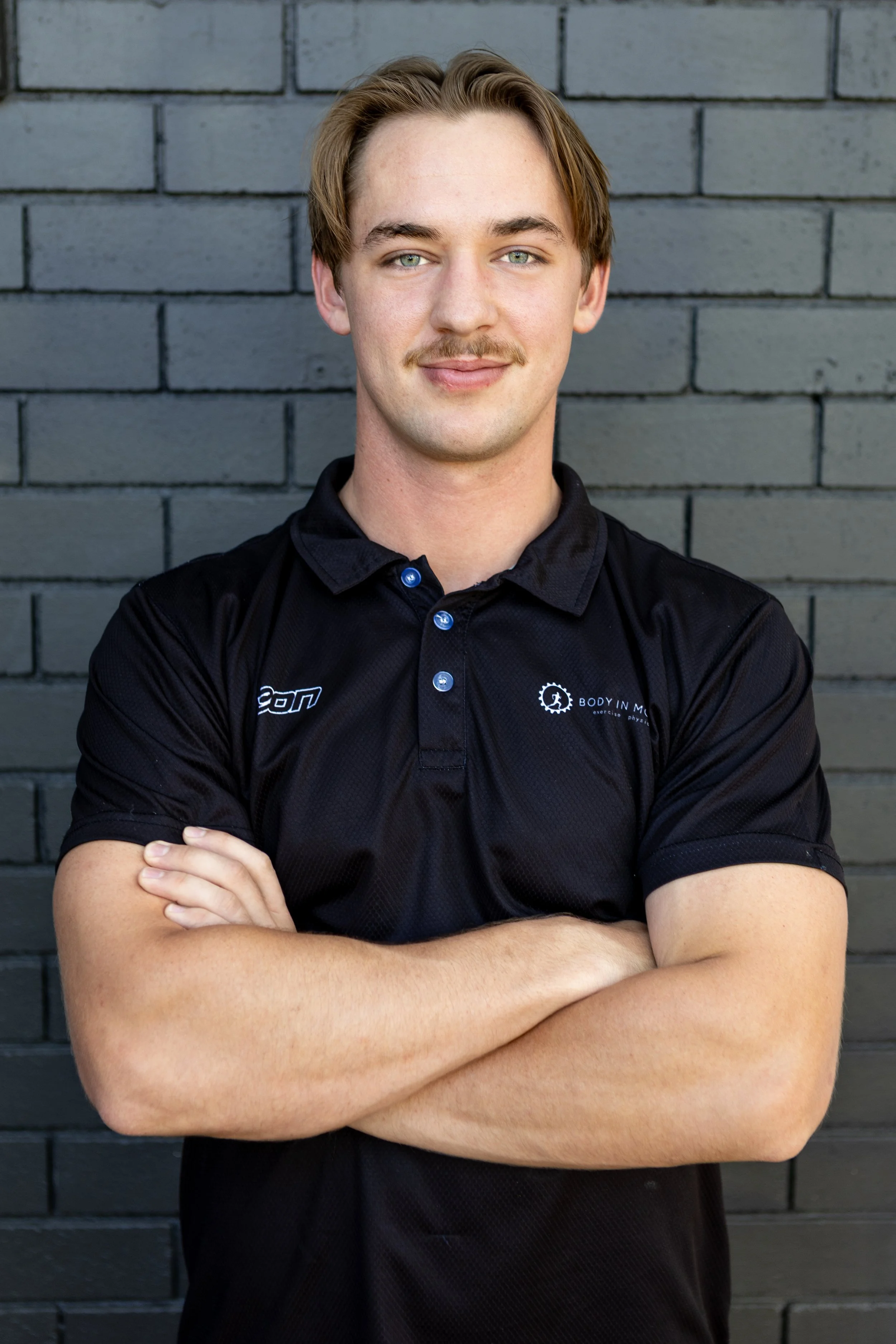 A young man with light skin, blonde hair, and a mustache, standing with arms crossed in front of a dark gray brick wall, wearing a black sports polo shirt with logos.