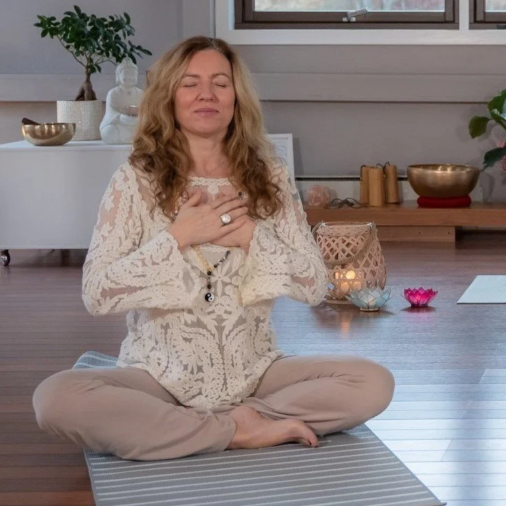 En kvinne med langt, blond hår sitter i lotusstilling på en yogamatte med øynene lukket, med hånden på brystet, inne i et rom med natur- og rostil. Det er lys fra et vindu bak henne.
