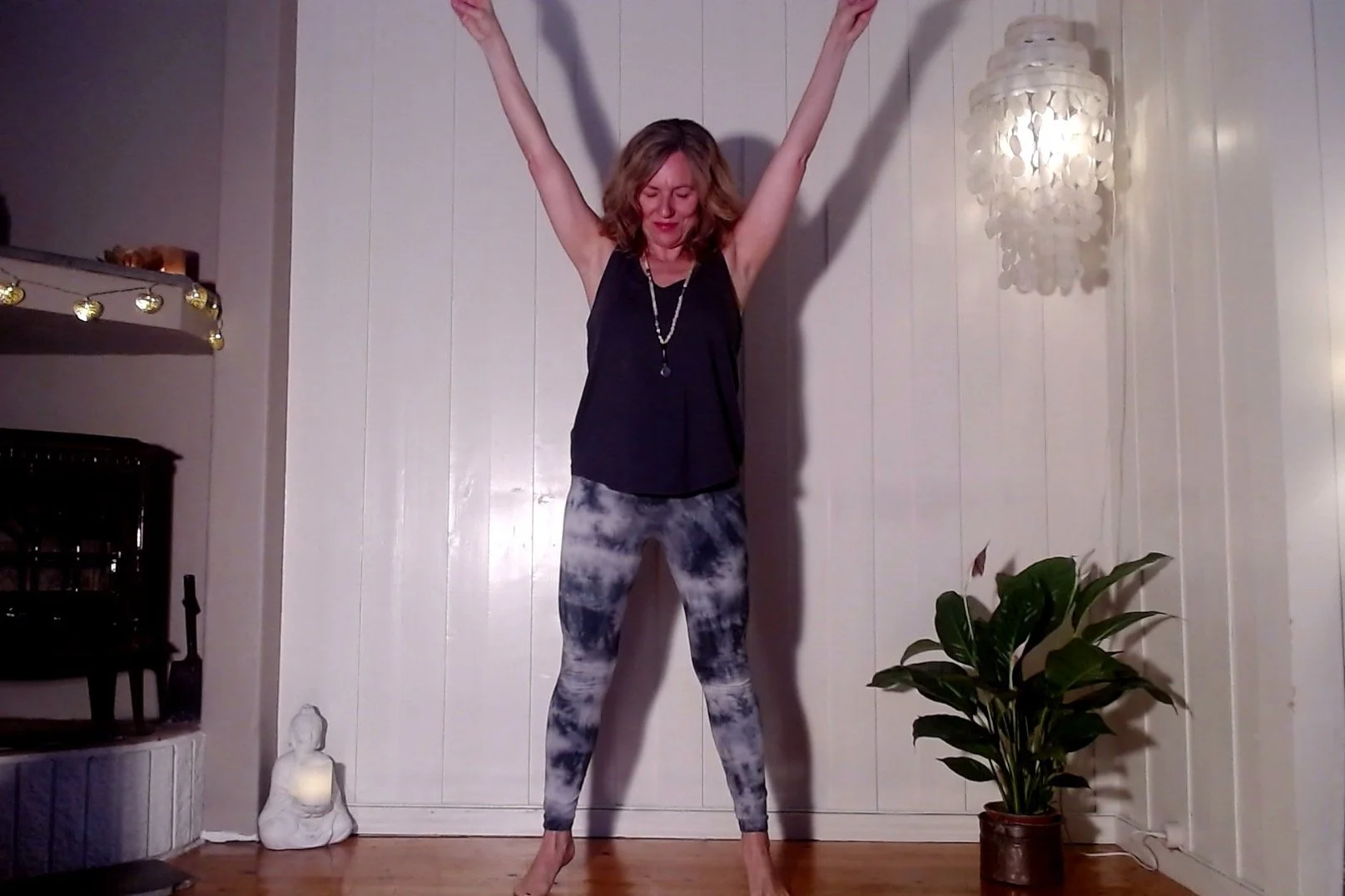 Kvinne står oppreist med armene hevet i en yoga-posisjon i et rom med lys vegg og gulv, omgitt av planter og dekorasjoner.