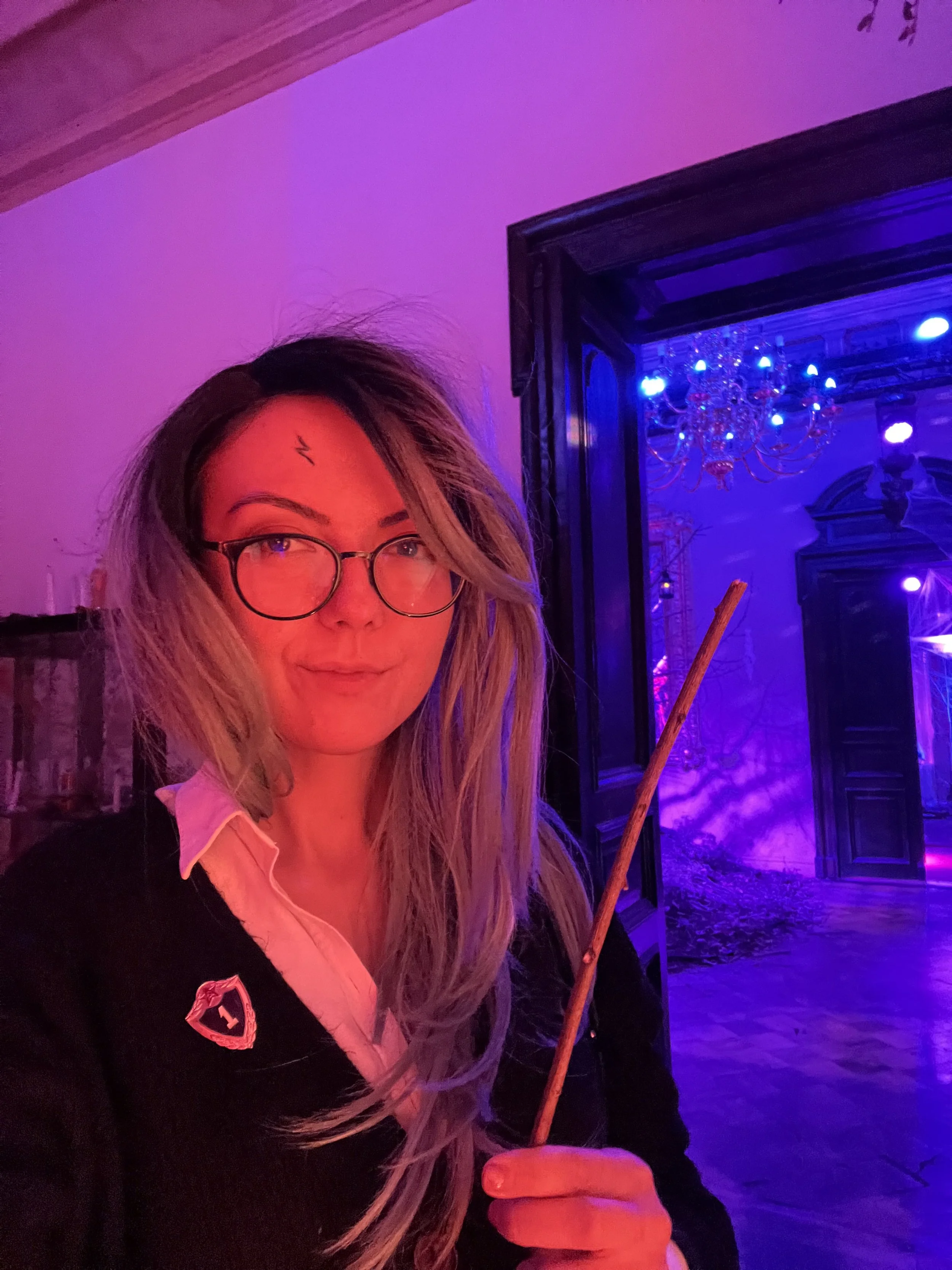 A woman with long blonde hair, glasses, and a small lightning bolt tattoo on her forehead, holding a stick, standing in a room with purple and blue lighting, a chandelier, and an ornate door.