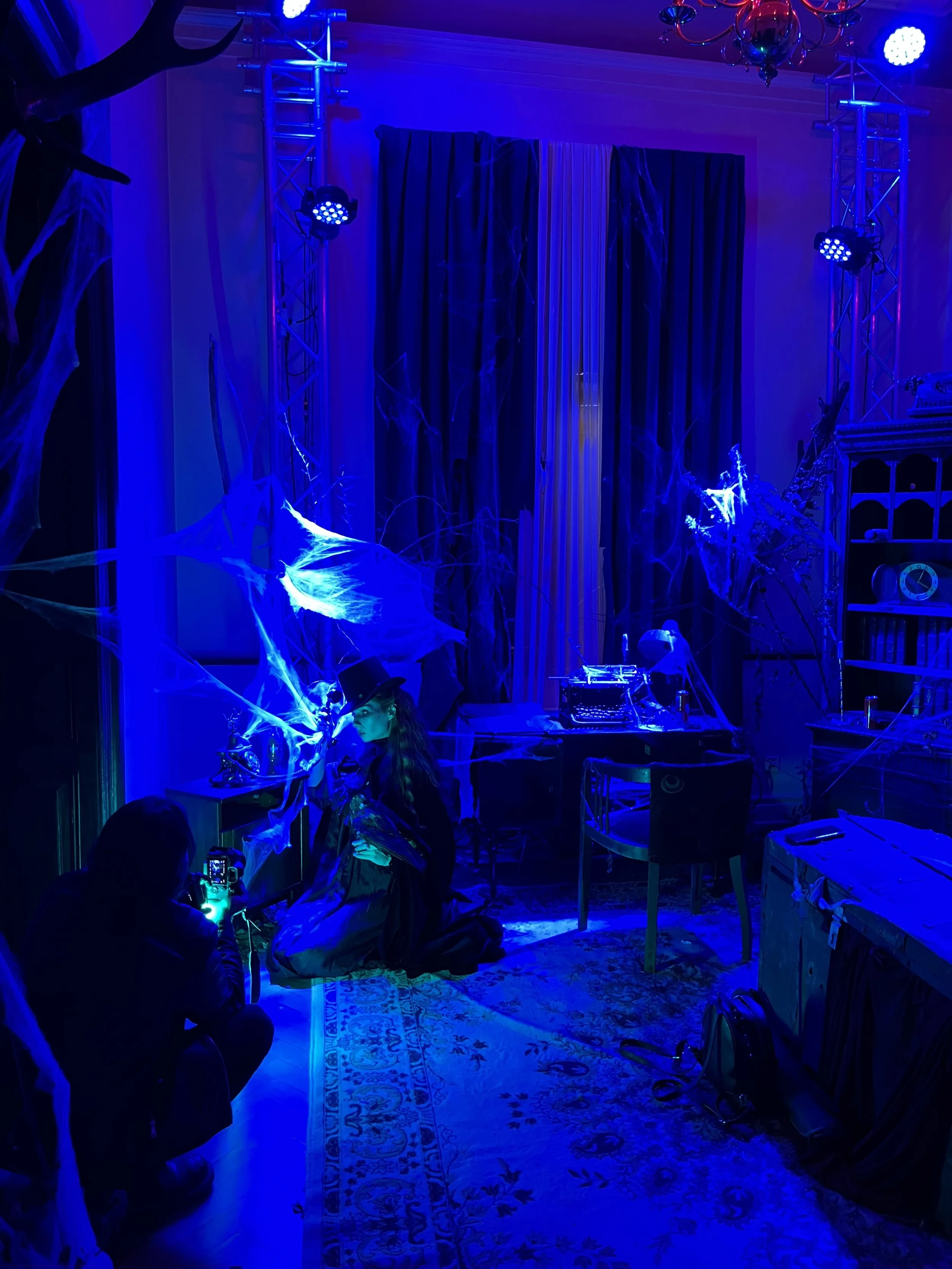Indoor room decorated for Halloween with blue lighting, fake cobwebs, and spooky decorations. A woman in a costume and a photographer are present in the room.