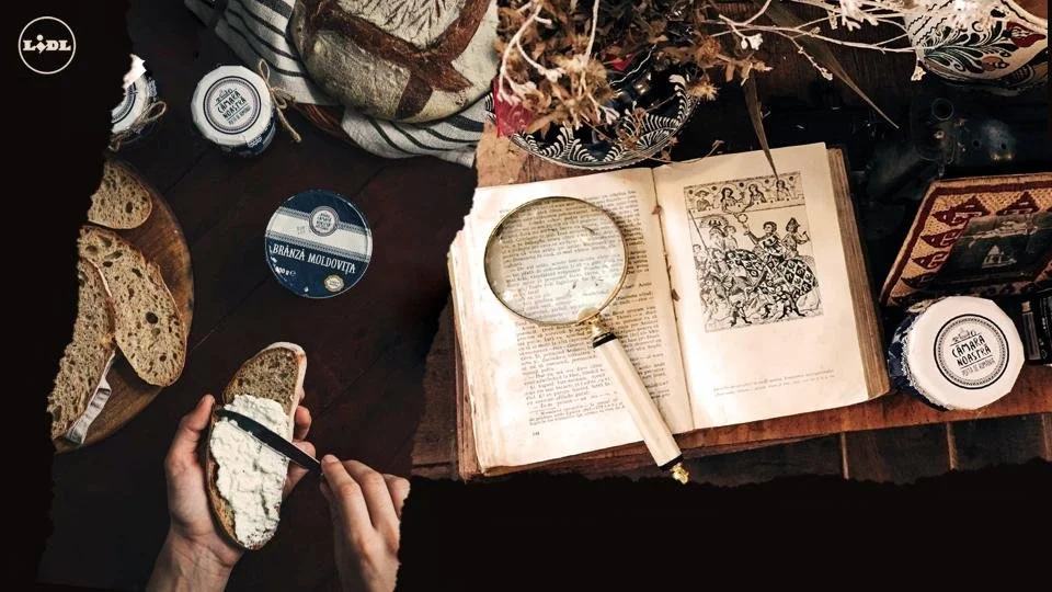 A wooden table with an open antique book, a magnifying glass resting on the book, a hand spreading cream or butter on a piece of bread, several jars and containers, some slices of bread, and decorative dried flowers.
