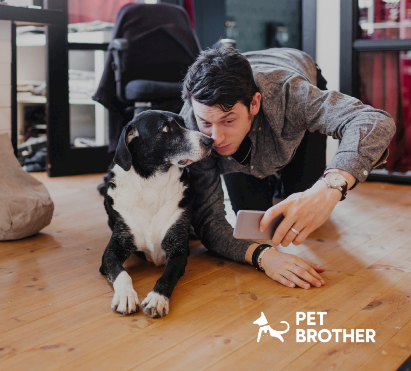 A young man lying on the wooden floor taking a selfie with a black and white dog. There is furniture and a backpack in the background. The logo for Pet Brother is in the lower right corner.