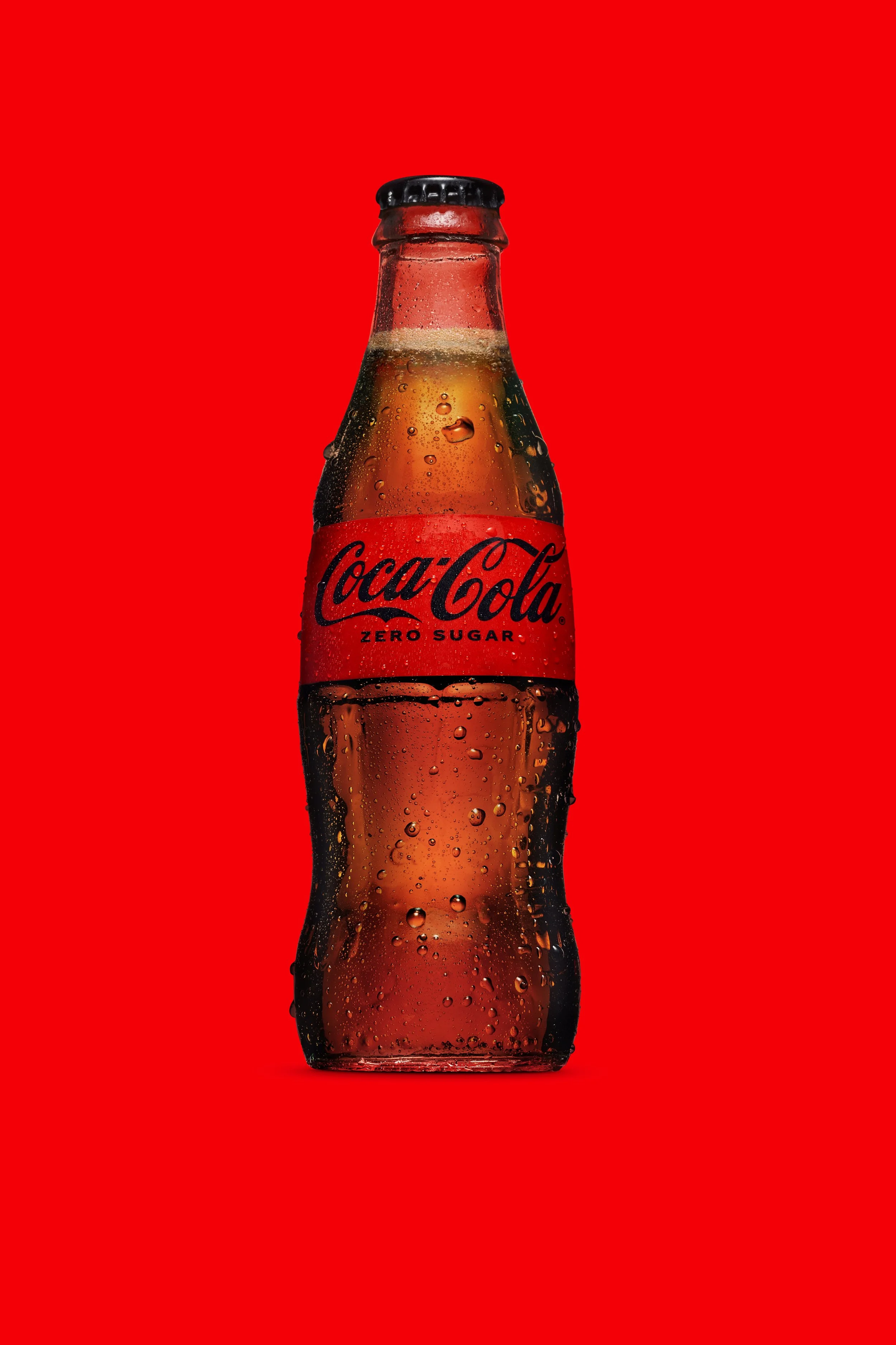 A glass Coca-Cola Zero Sugar bottle with condensation droplets on a red background.