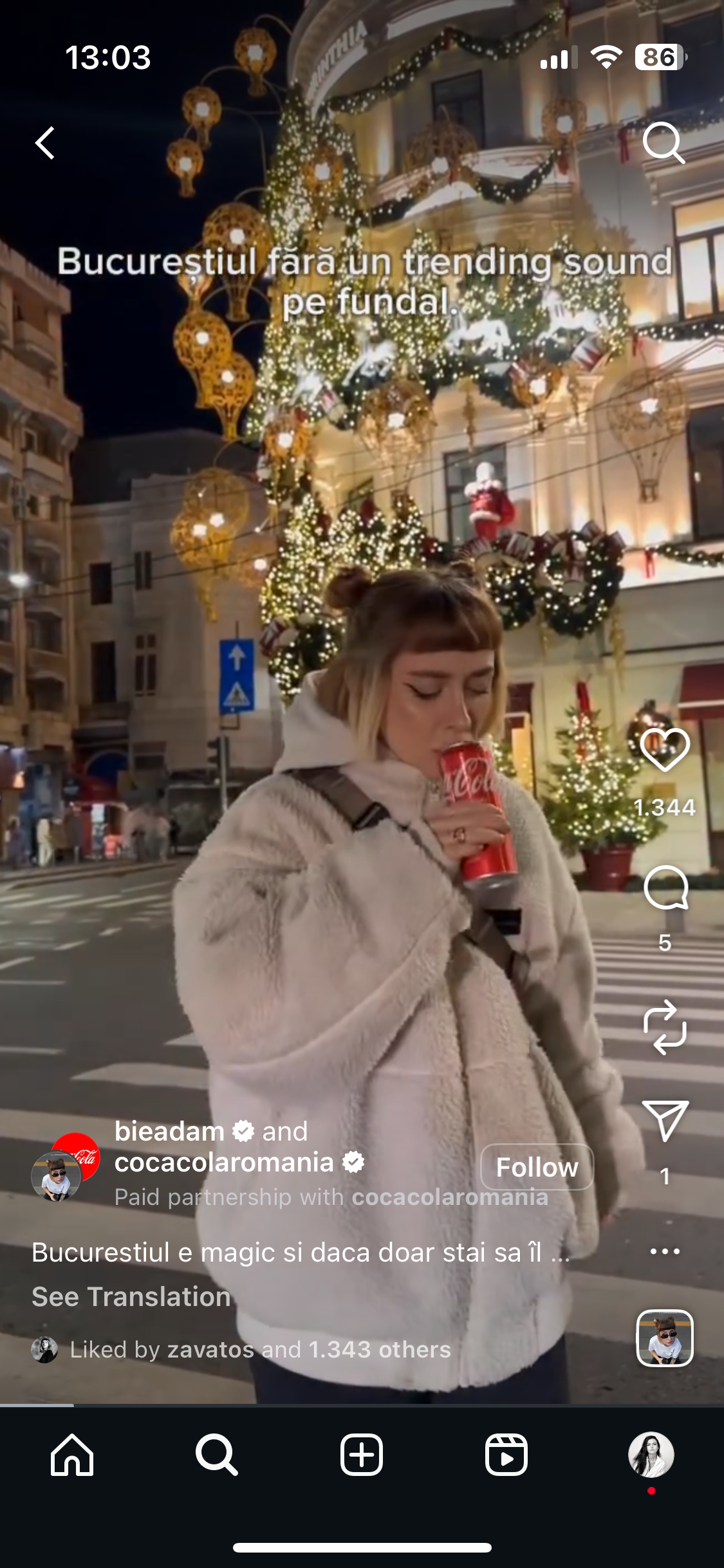 A young woman with light brown hair styled in buns, wearing a white fuzzy jacket and holding a Coca-Cola can, stands on a city street at night in front of a decorated building with Christmas lights, wreaths, and ornaments with a large Christmas tree in the background.