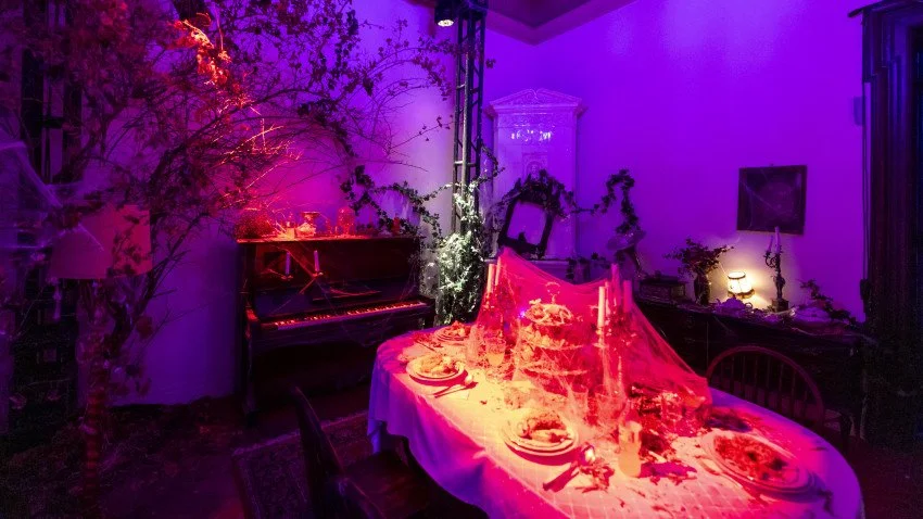 A dining room illuminated with purple and pink lighting, featuring a table set with plates and glasses, a vintage portrait with candles, a piano in the background, and Halloween or spooky decorations like spider webs and branches.