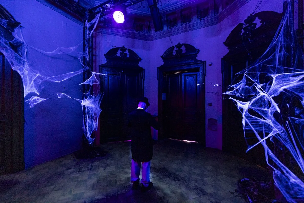 Person dressed as a detective with a hat and trench coat stands in a dark room decorated with glowing spider webs illuminated by purple light.