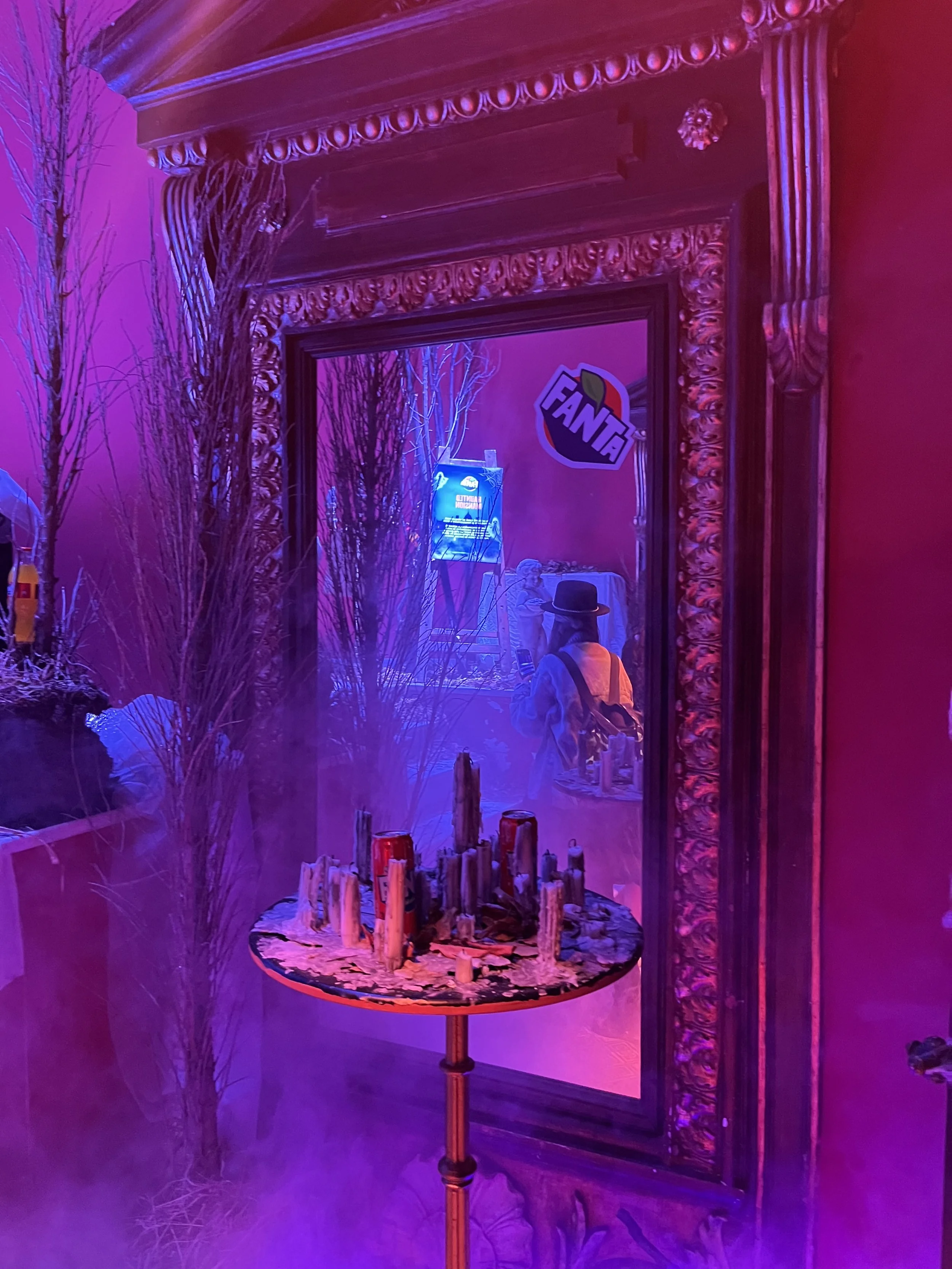 Reflection of a person in a mirror with a decorative frame, neon lights, a table with metallic objects, and a 'Fanta' sign in a dimly lit room with purple and blue lighting.