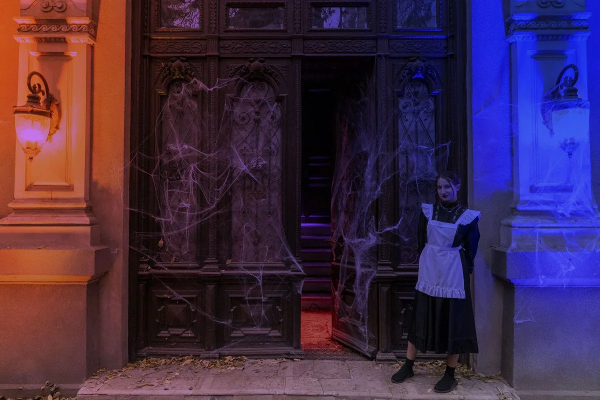A woman dressed as a maid in a vintage black dress with a white apron standing outside a large wooden gate decorated with cobwebs, illuminated by purple and orange lighting, with cobwebs in the scene and fallen leaves on the ground.